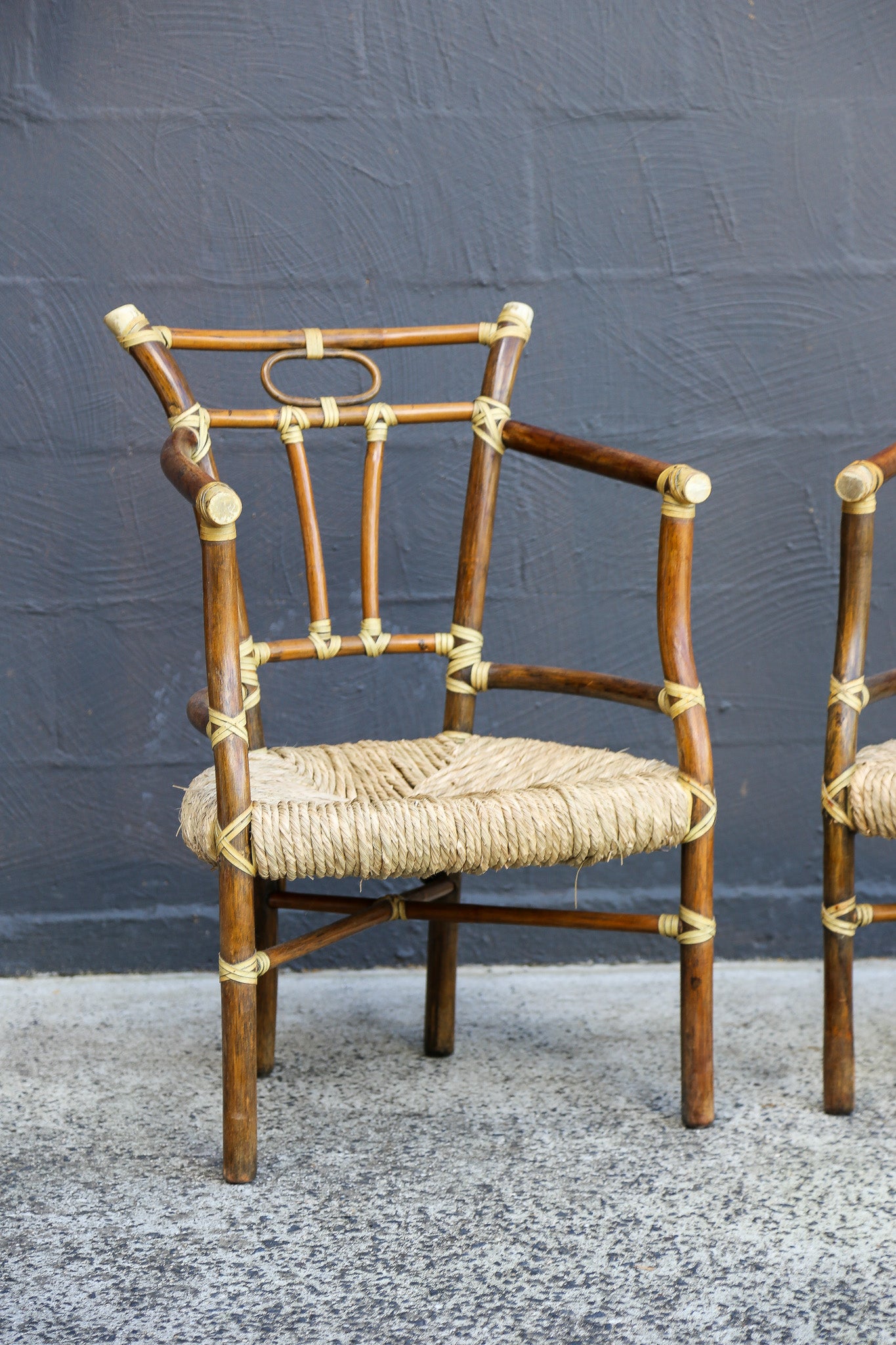 A mid-century modern armchair with a dark brown bamboo frame, rope-wrapped seat and armrests, and brass hardware, set against a dark gray textured wall.
