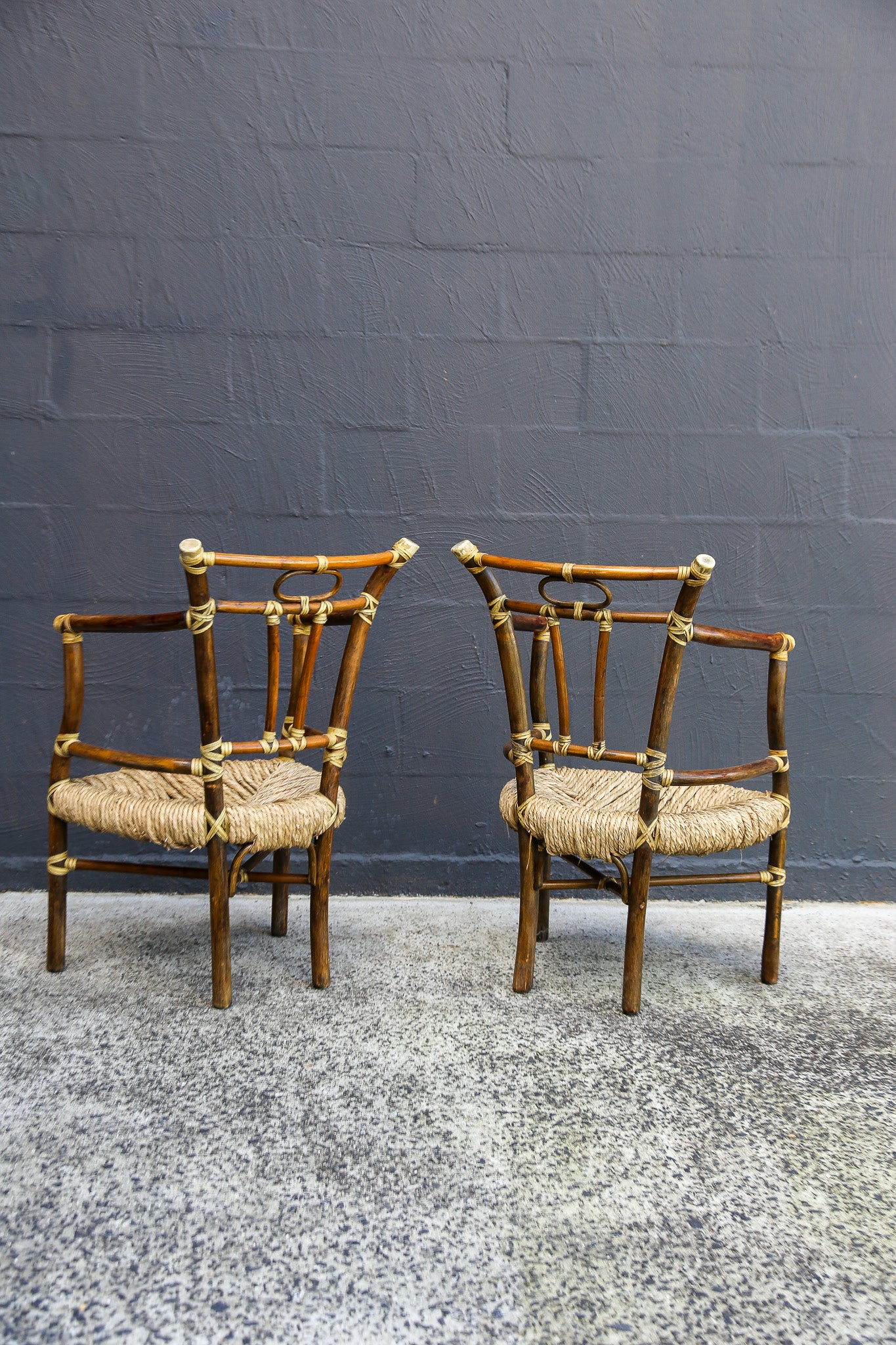 Two vintage bamboo armchairs with woven rattan seats, positioned side-by-side against a dark gray brick wall and speckled gray floor.