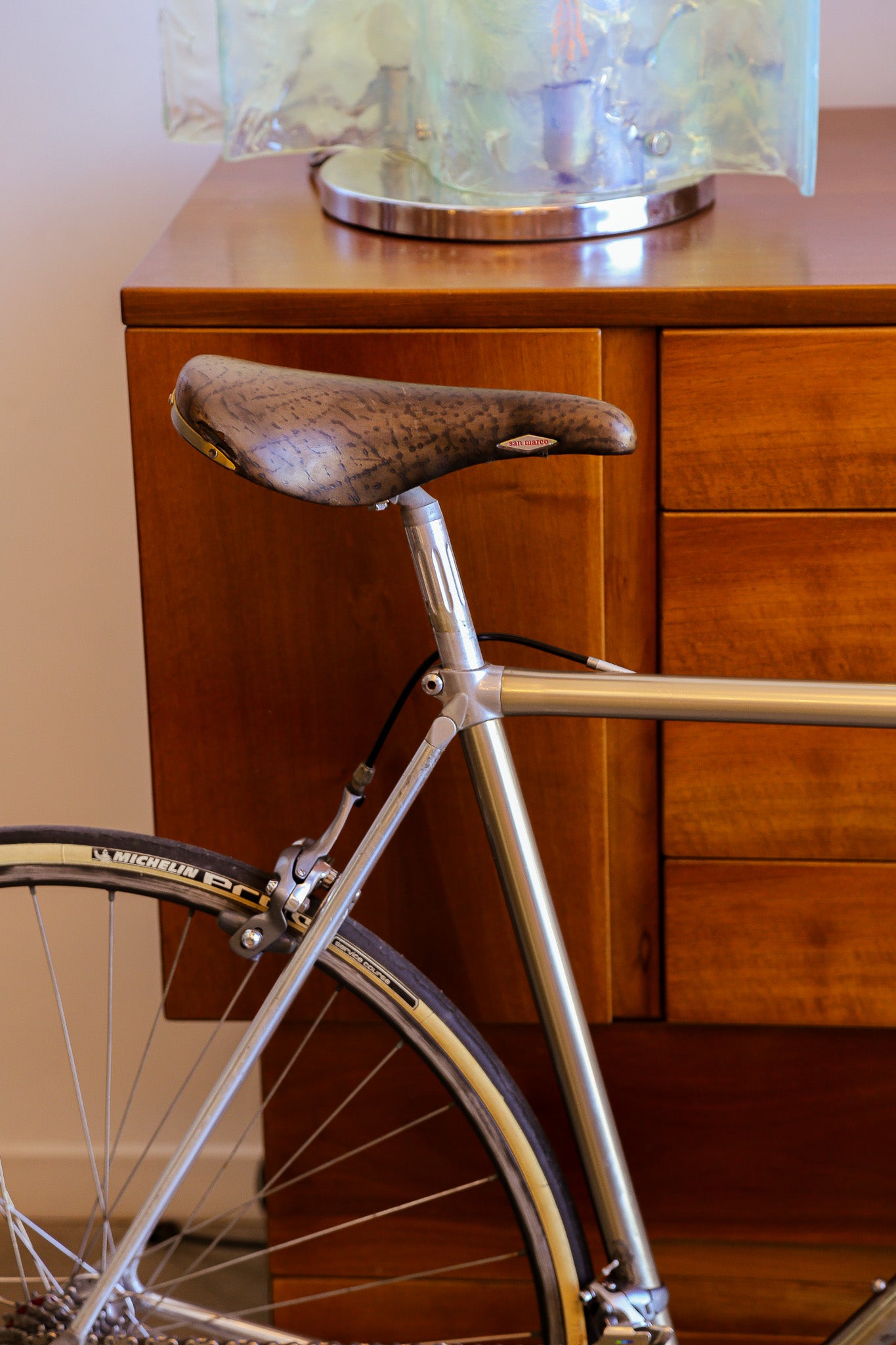 A close-up of a vintage-style bicycle frame with a brown leather saddle and a silver metal frame, positioned in front of a wooden cabinet.