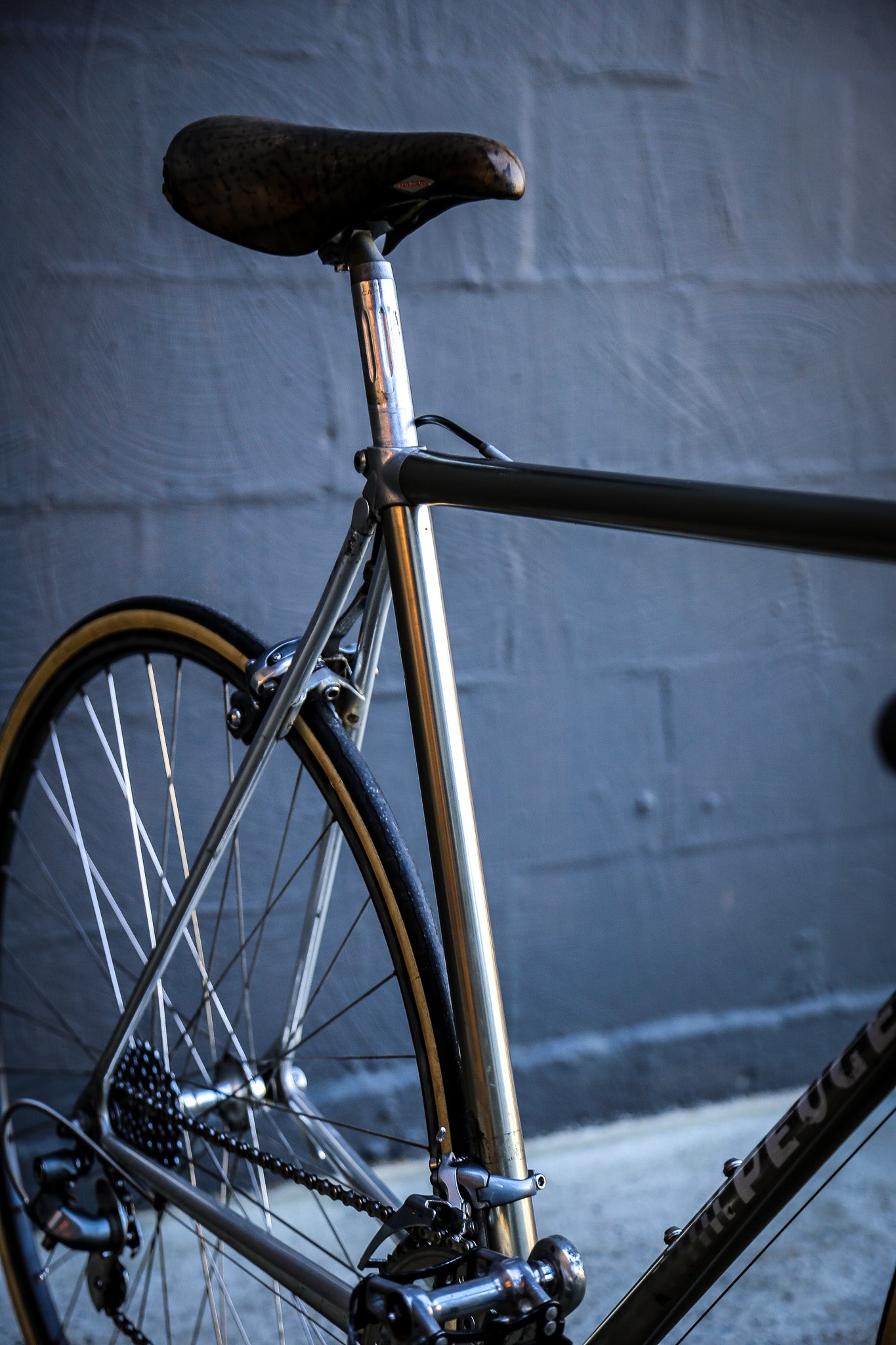 Close-up of a vintage-style road bicycle frame in metallic silver, showing the seat post, rear wheel, and part of the down tube with visible 'Devyne' branding, set against a gray brick wall.