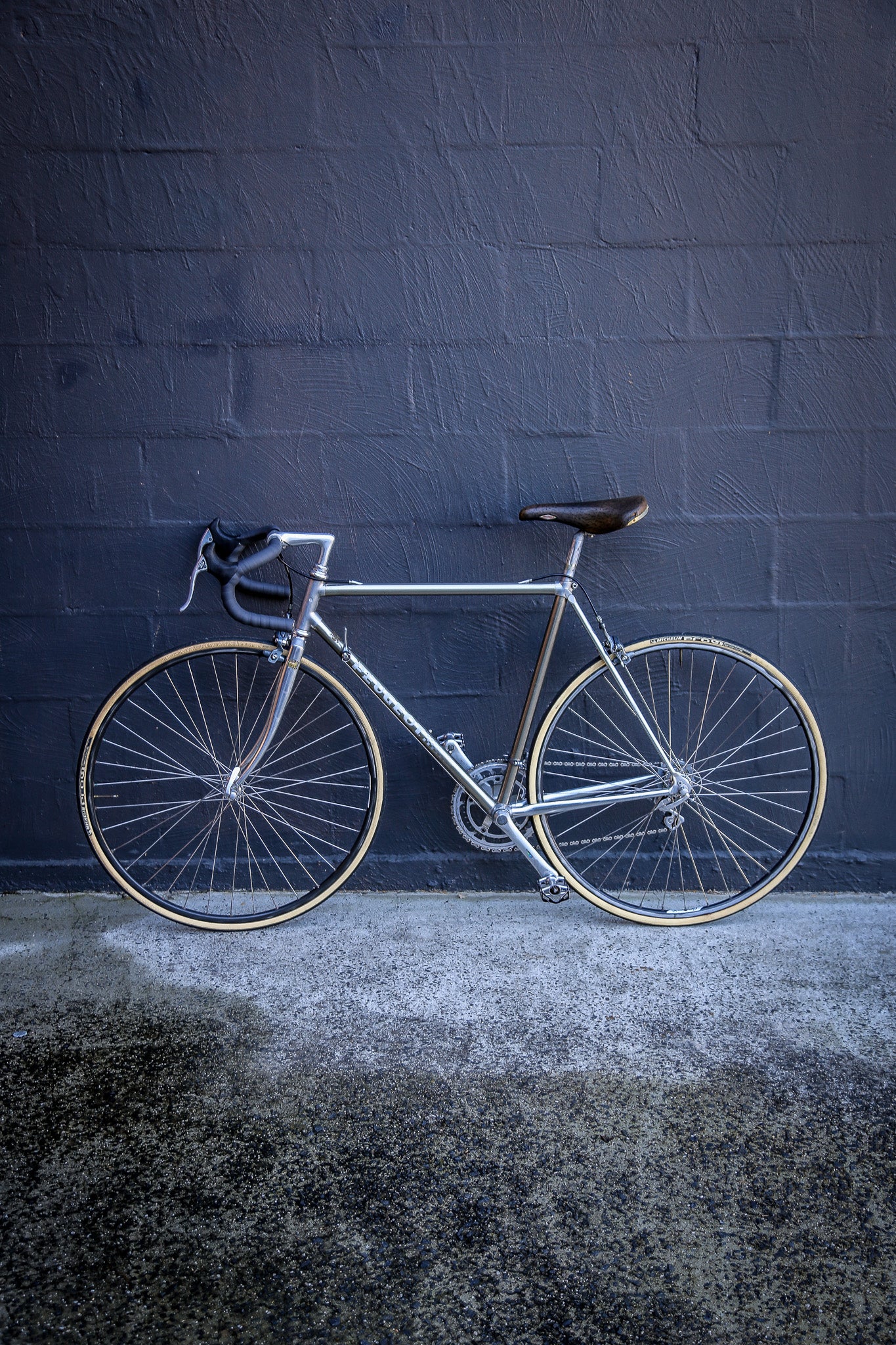 A silver road bicycle with thin tires and a dark saddle is parked against a dark gray painted brick wall on a textured concrete ground.