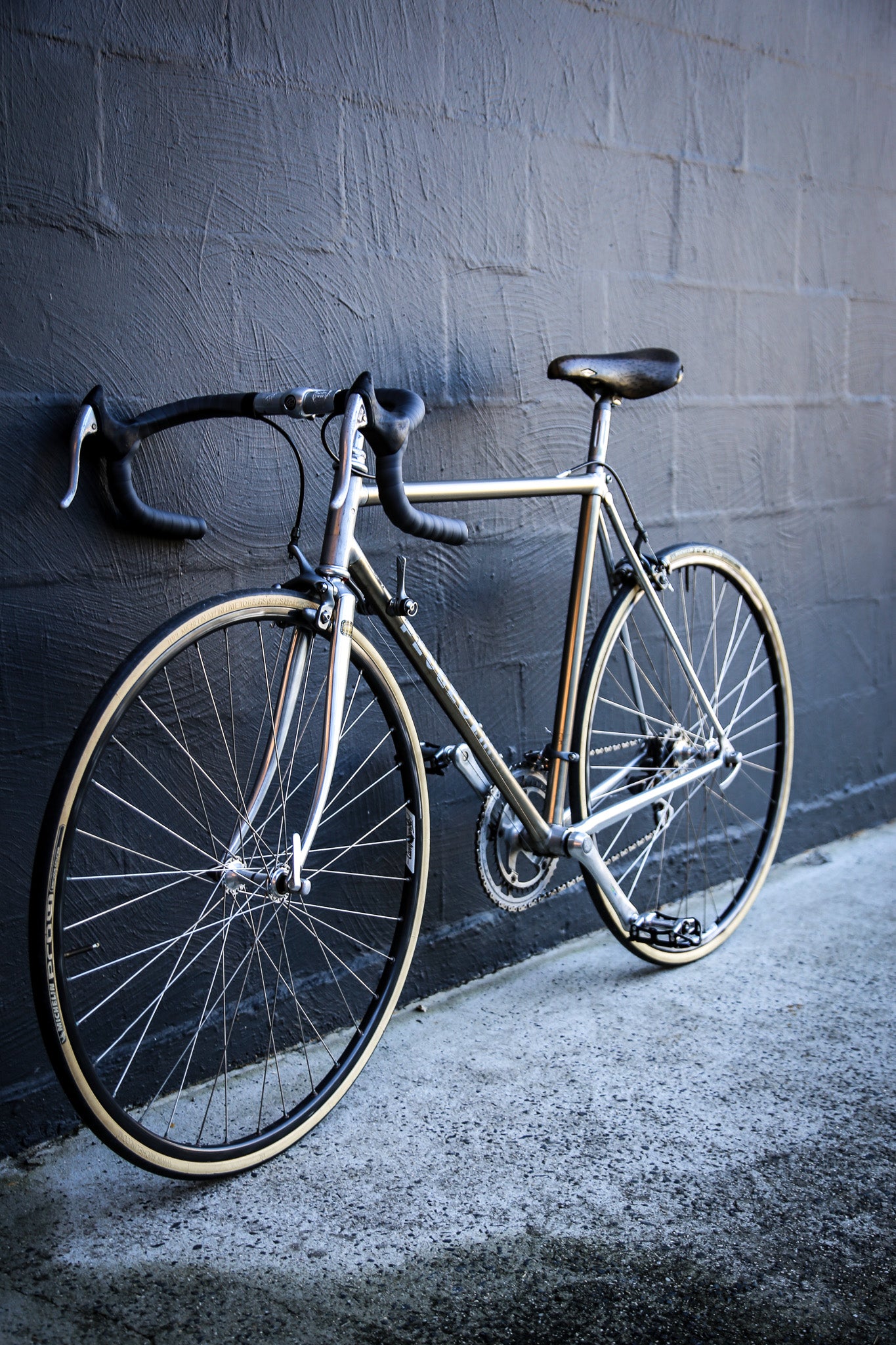 A vintage-style road bicycle with a silver frame, black drop handlebars, and black saddle, leaning against a dark gray brick wall on a concrete ground.