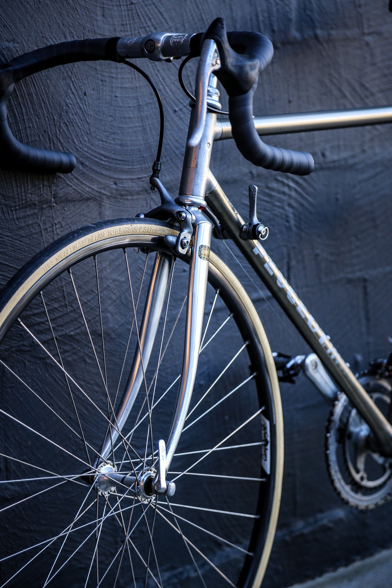 Close-up of a vintage-style road bicycle with a metallic silver frame, black drop handlebars, and a thin black tire, leaning against a dark textured wall.