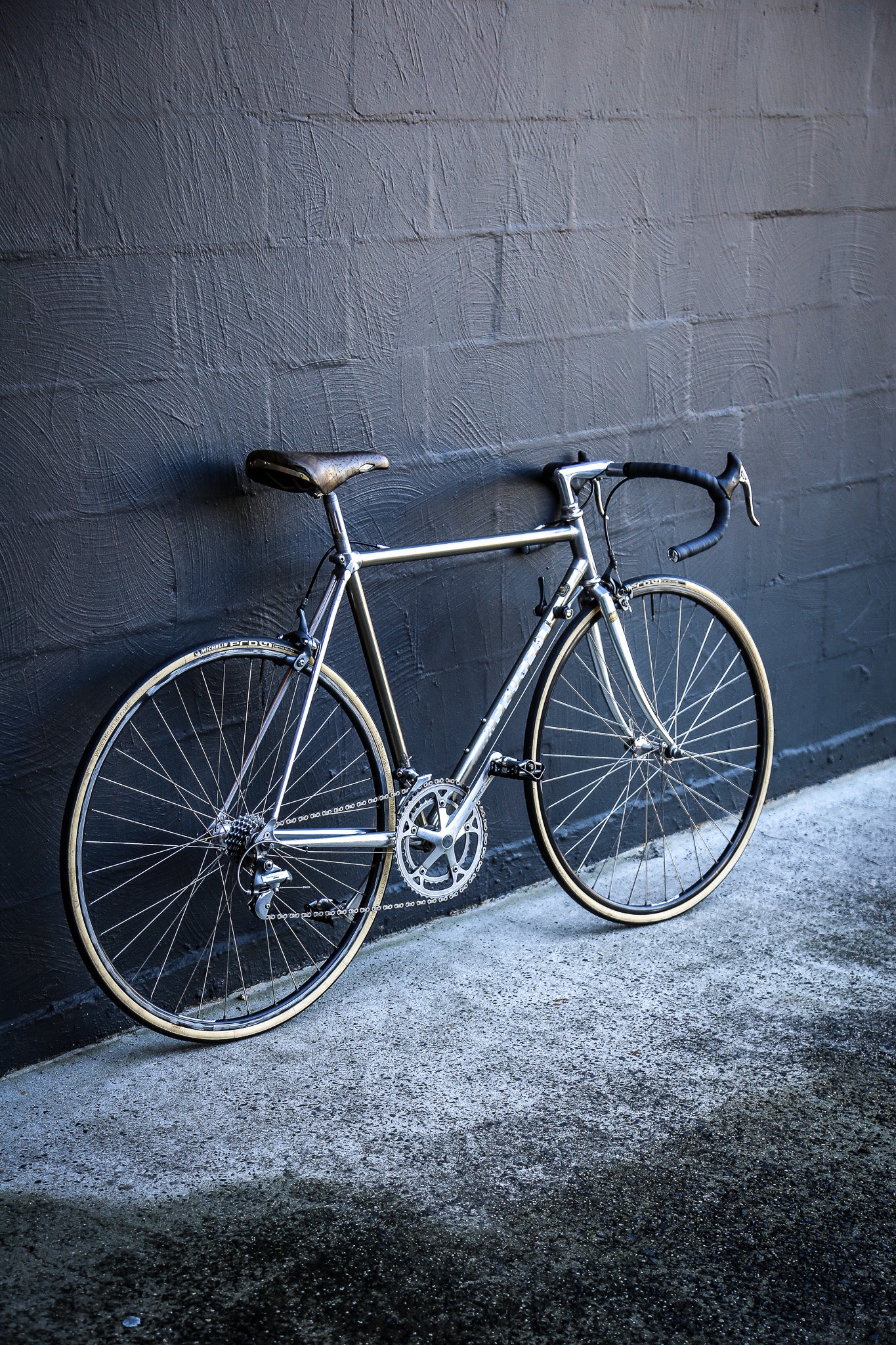 A silver road bicycle with black drop handlebars and a black saddle, leaning against a dark gray brick wall on a concrete ground.