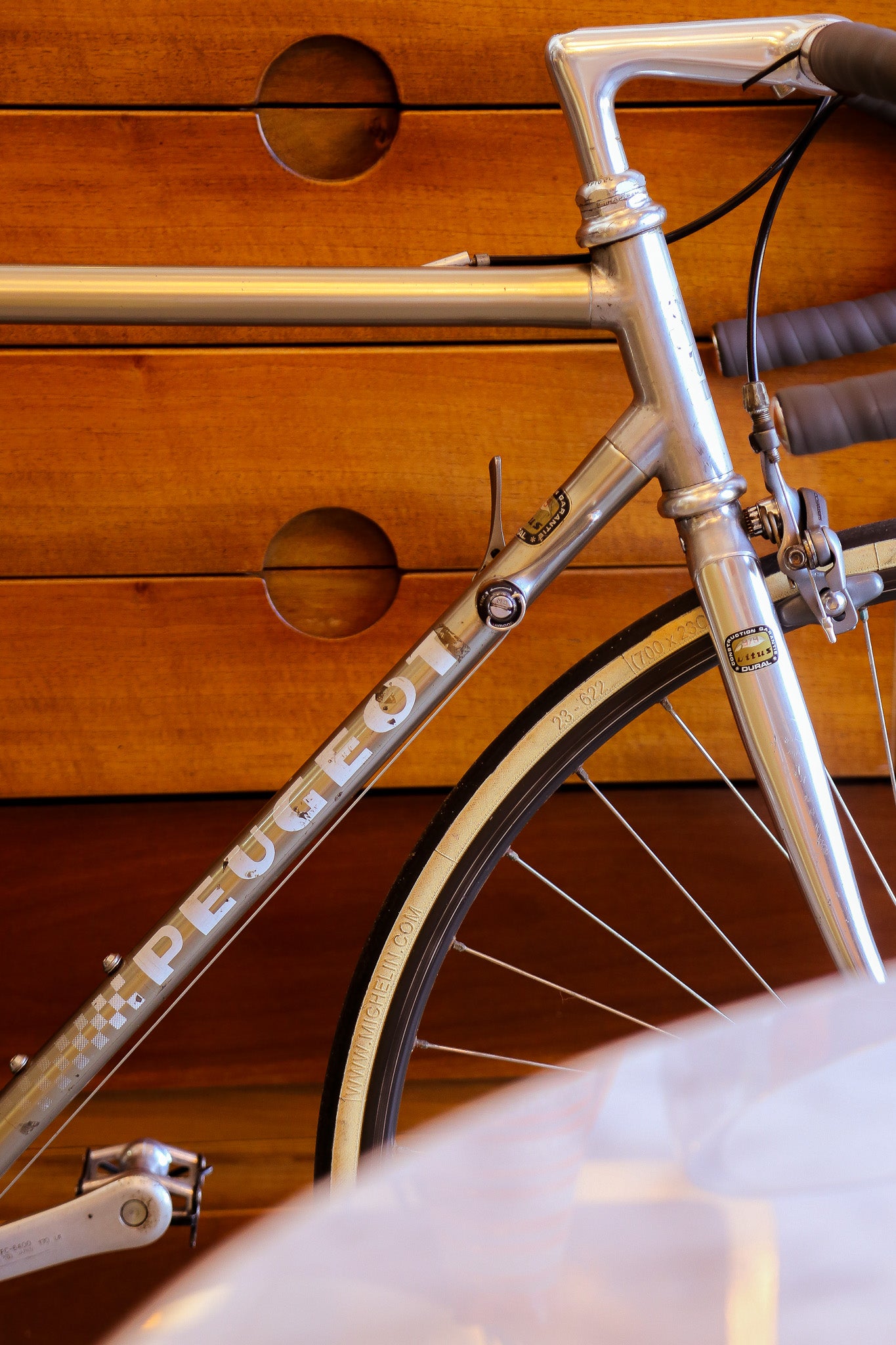 Close-up of a vintage silver Peugeot road bicycle frame with the brand name clearly visible on the down tube, set against a warm wooden background.