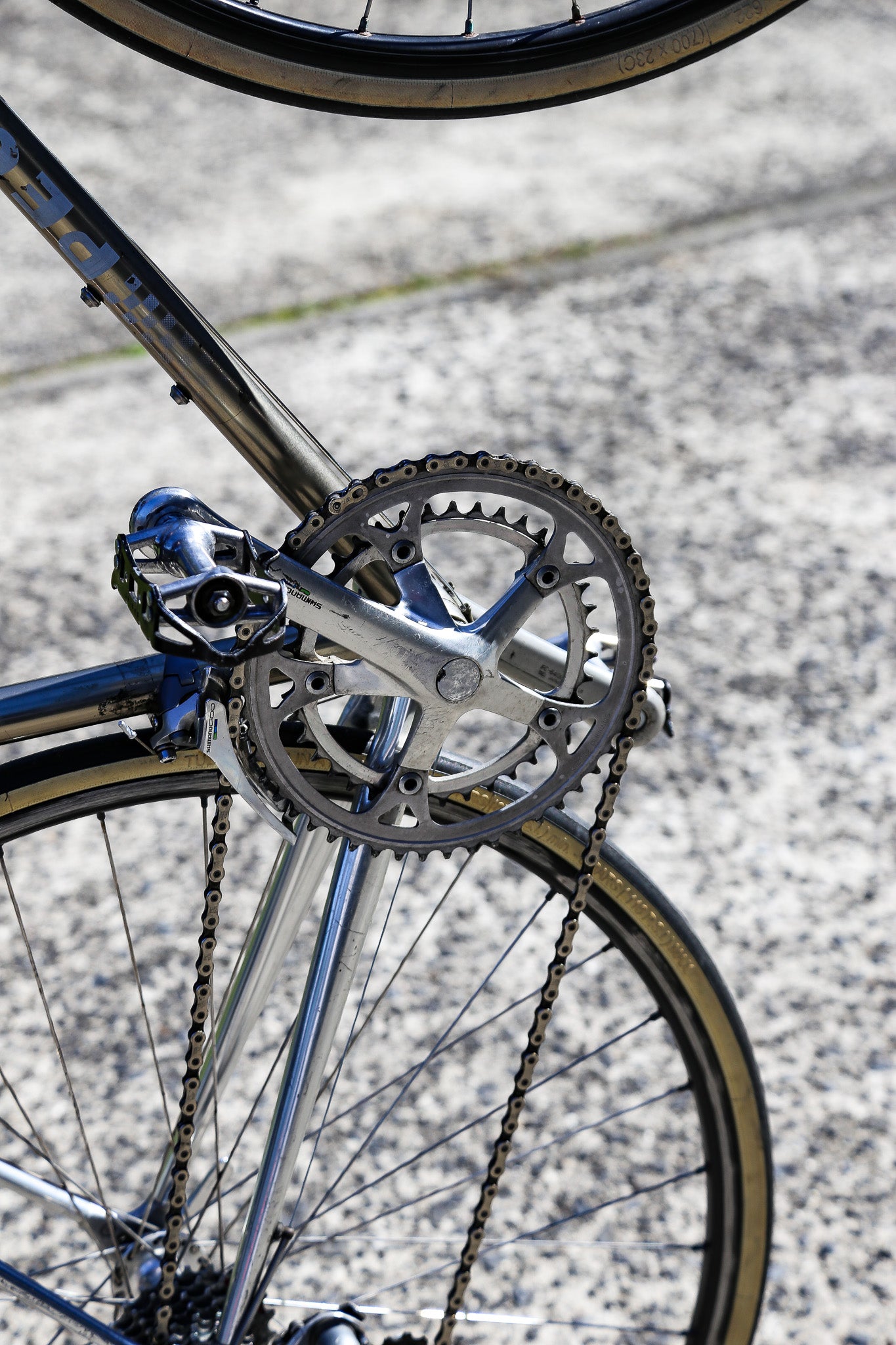 Close-up of a bicycle crankset with a silver chainring, chain, and rear wheel on a paved road background.