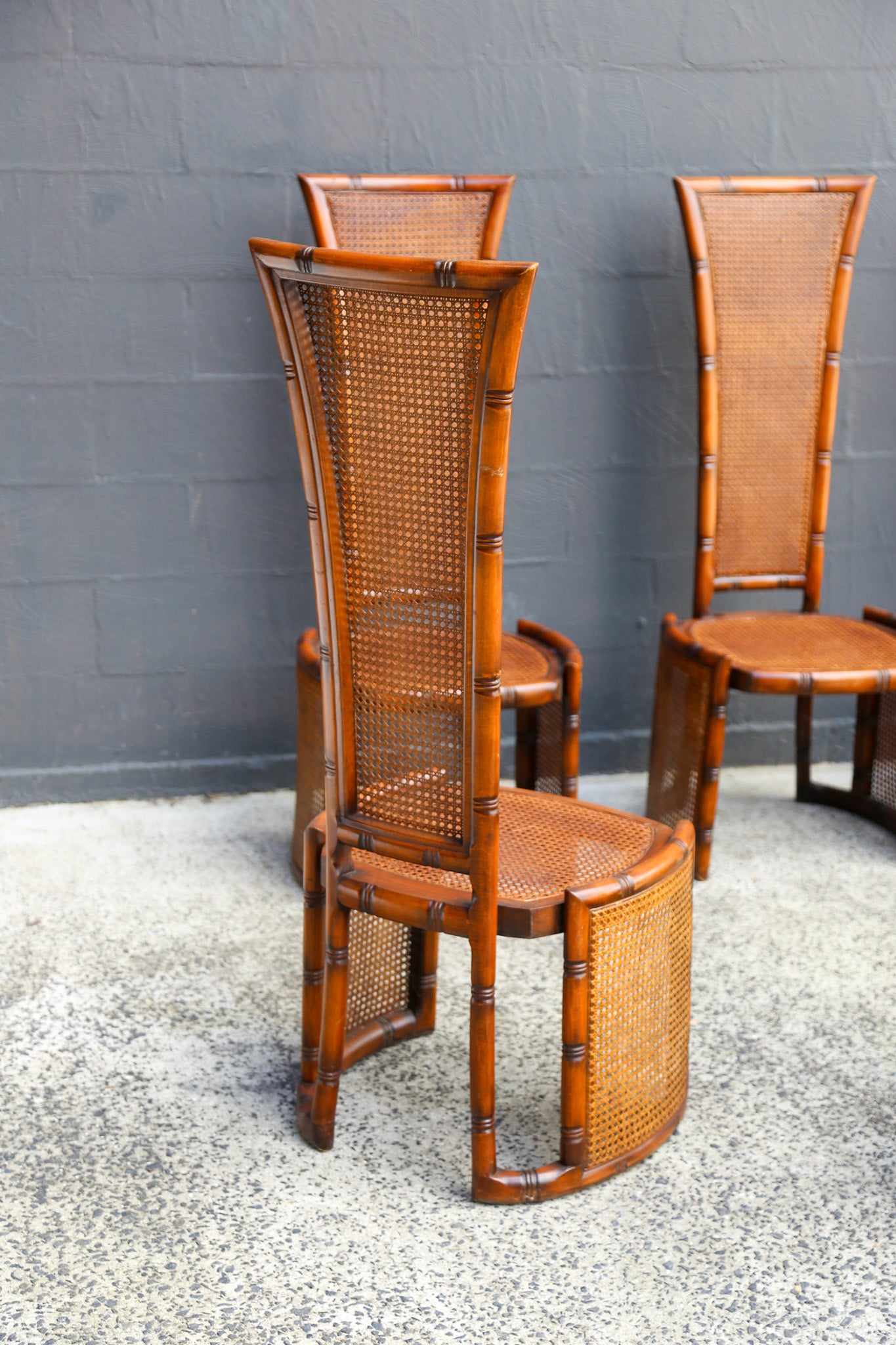Three mid-century modern bamboo dining chairs with woven cane backs and seats, set against a dark gray brick wall and speckled concrete floor.