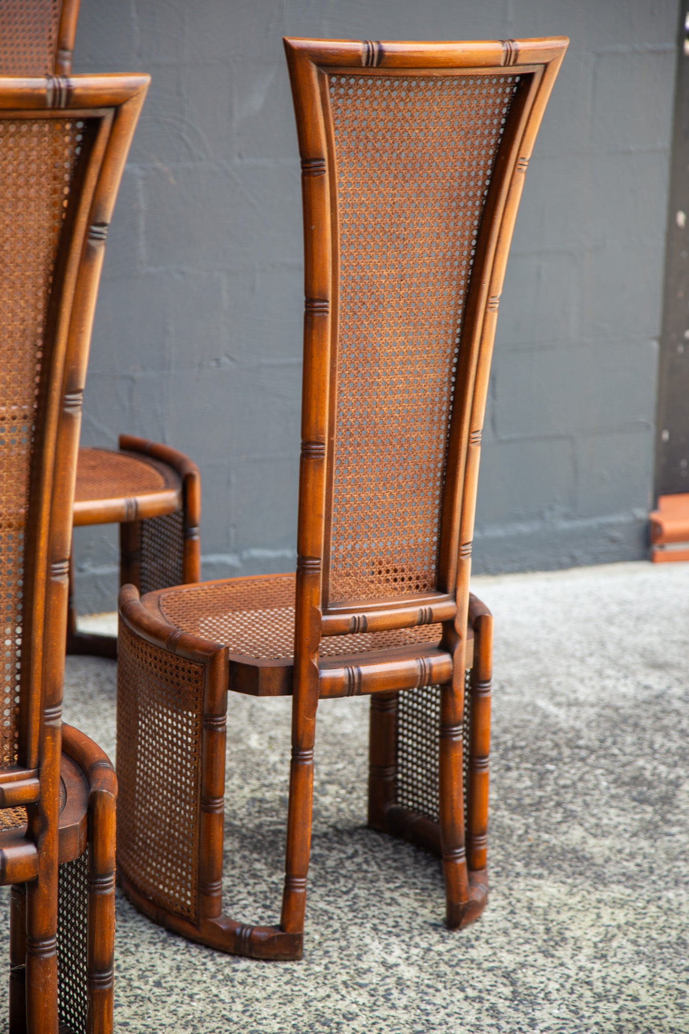 A tall bamboo-style dining chair with woven cane back and seat, shown in a real setting against a gray wall and speckled floor.
