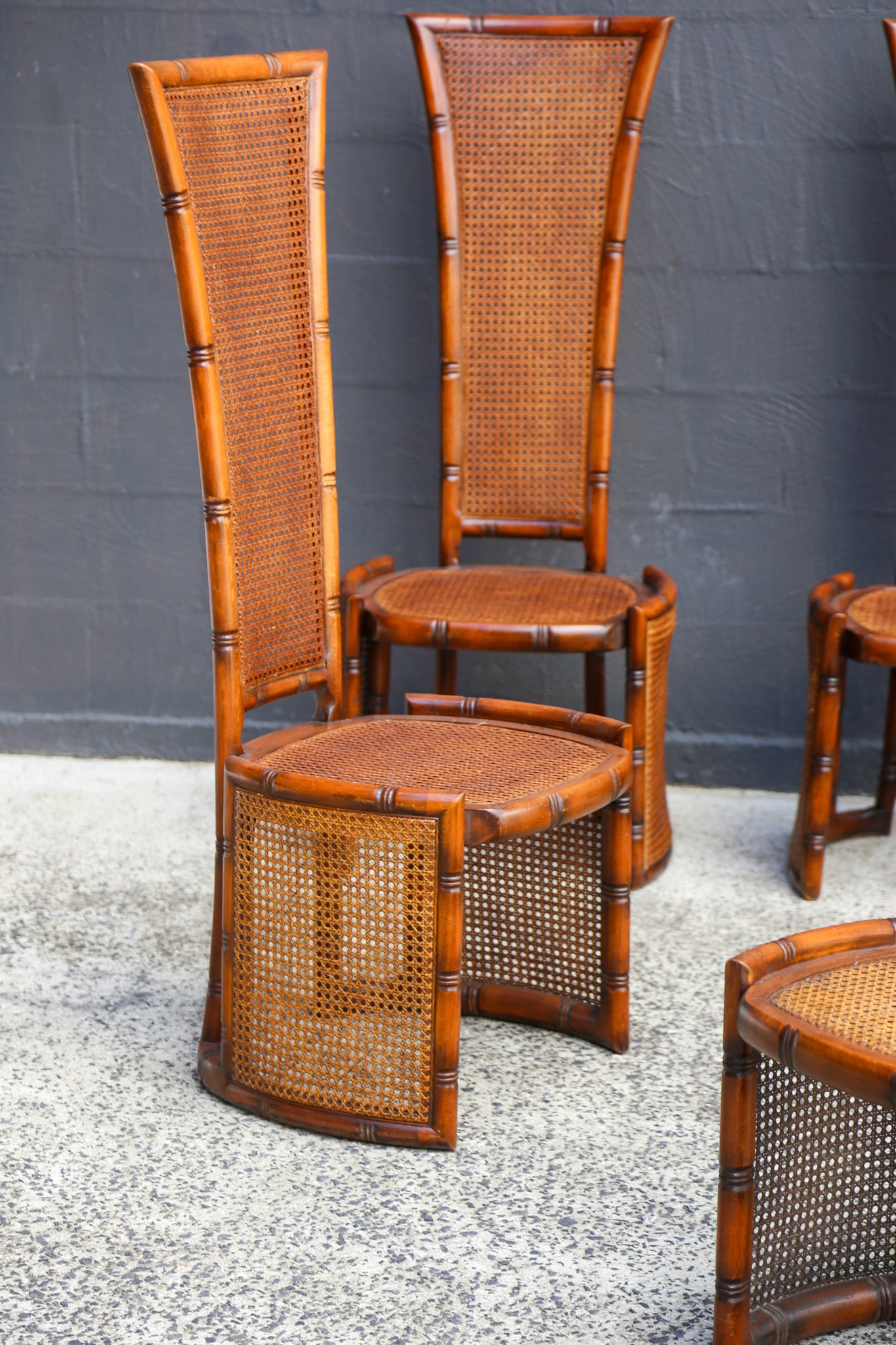 A set of mid-century modern bamboo and cane chairs with tall woven backs and round cane-slat seats, shown against a dark gray brick wall and speckled concrete floor.