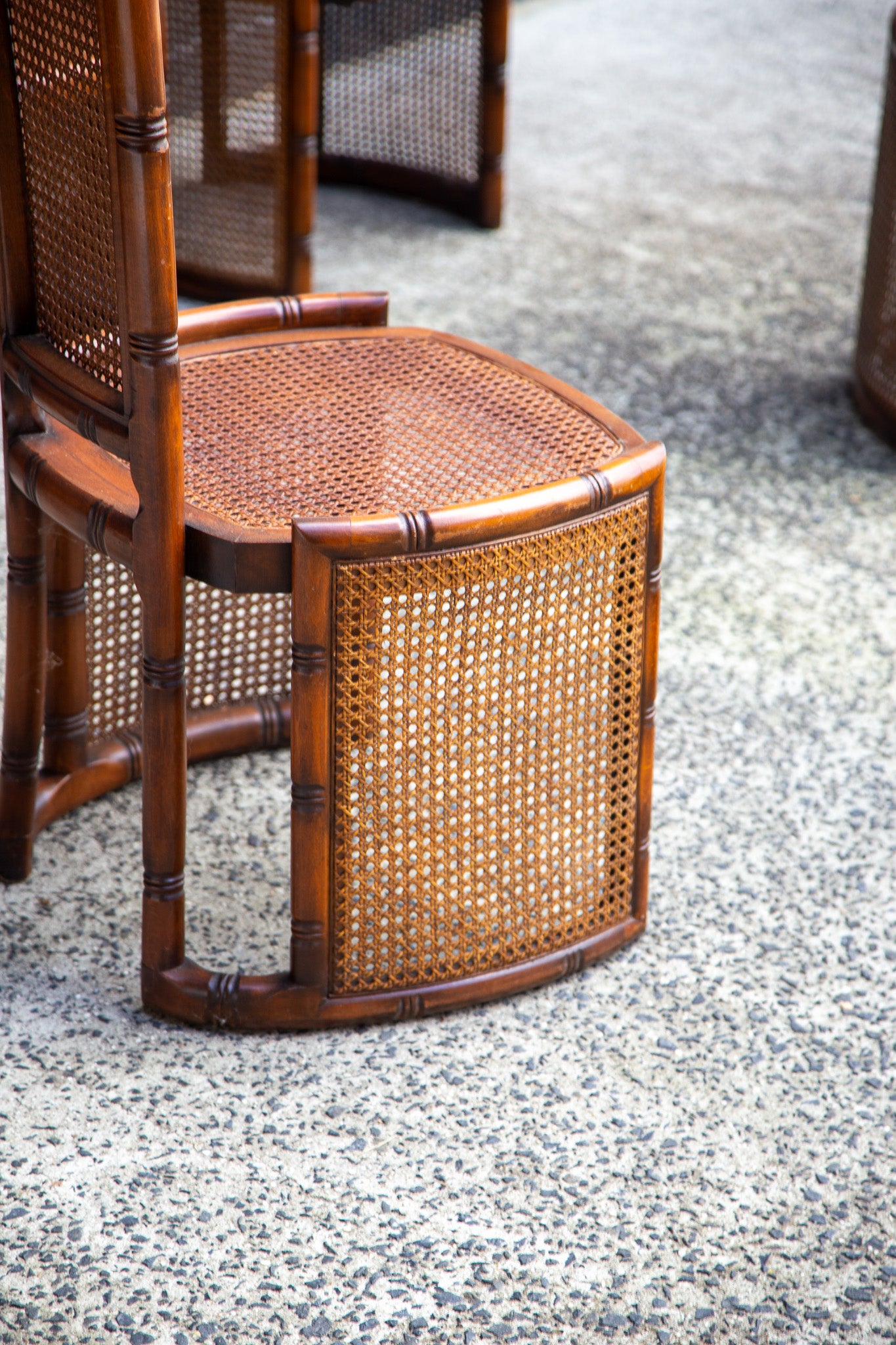 A close-up of a wooden dining chair with a woven cane seat and back, shown on a speckled concrete floor with other matching chairs blurred in the background.