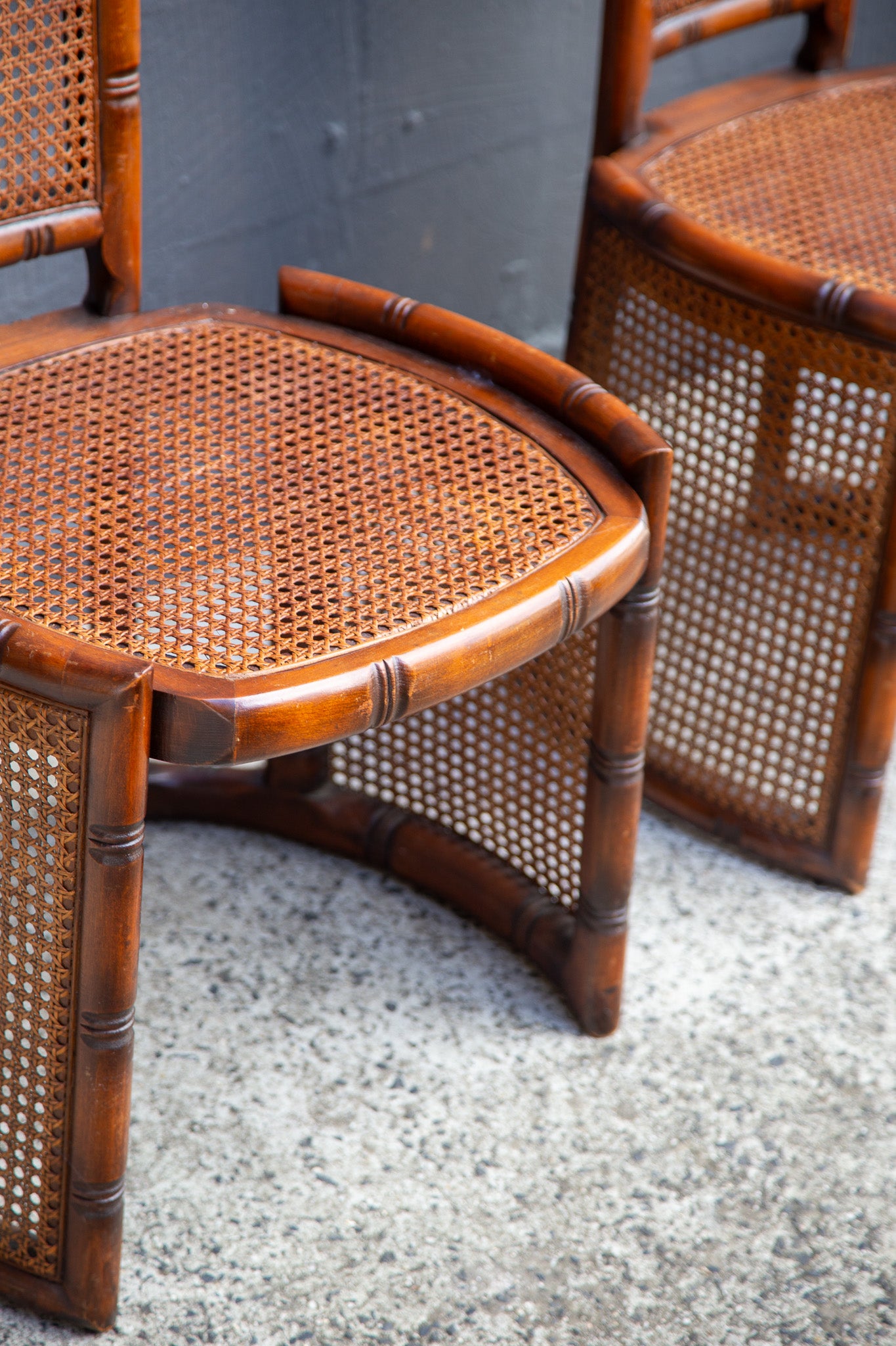 A close-up of a curved bamboo armchair with a woven cane seat and back, shown alongside another matching chair on a speckled stone floor against a dark gray wall.