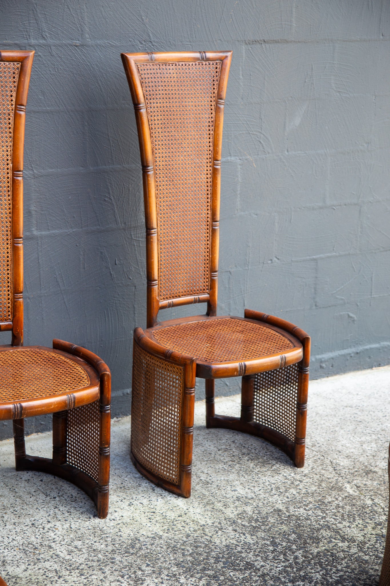 A mid-century modern wooden and cane dining chair with a tall slatted back and curved base, shown next to another matching chair against a gray brick wall.