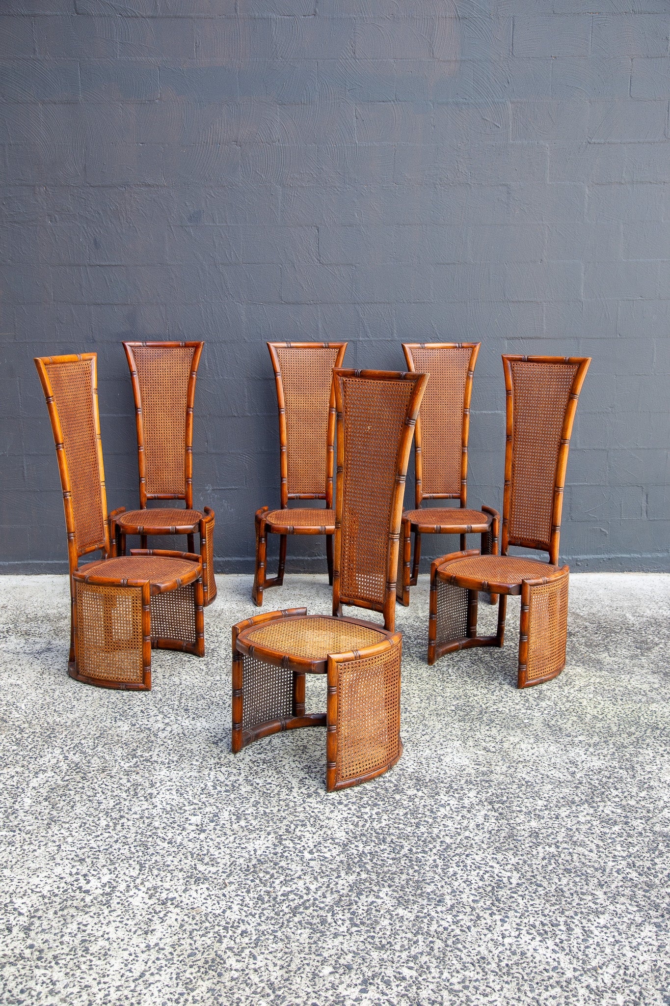 A set of six mid-century modern rattan dining chairs and two matching side tables, featuring tall woven cane backs and curved bamboo frames, arranged on a speckled concrete floor against a dark gray brick wall.