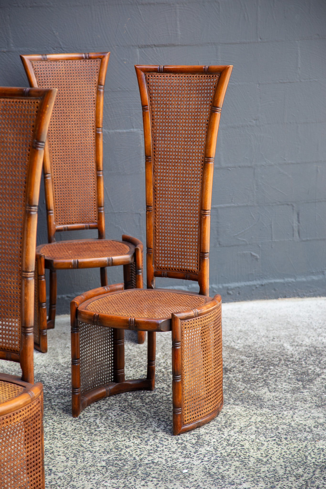 A set of mid-century modern rattan and bamboo chairs with tall cane backs and curved bamboo frames, arranged on a speckled concrete floor against a gray brick wall.
