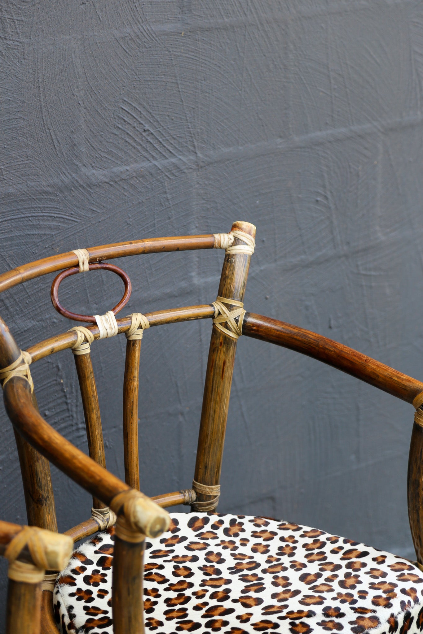 A close-up of a rattan or bamboo chair with a leopard-print upholstered seat cushion, set against a textured dark gray wall.