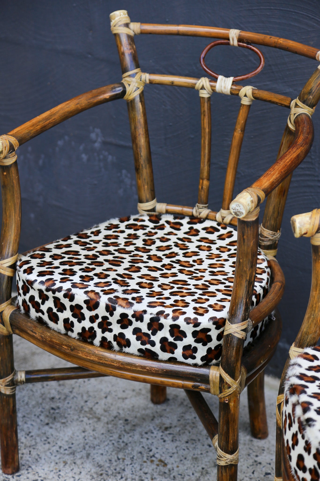 A close-up of a rattan armchair with a leopard-print upholstered seat cushion, set against a dark textured wall.