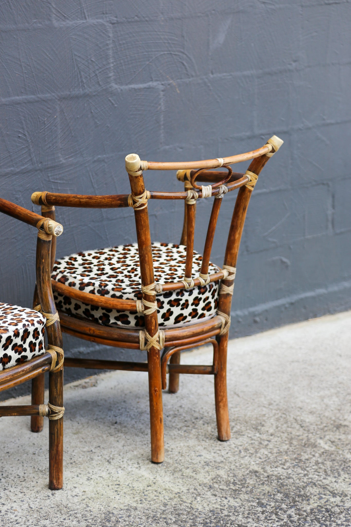 A rustic bamboo chair with a leopard-print cushion, positioned outdoors against a dark gray painted brick wall on a concrete floor.
