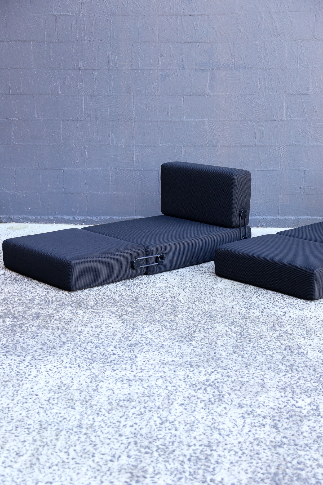 A black modular outdoor seating set consisting of a chaise lounge and two matching ottomans, positioned on a textured gray concrete surface against a gray painted brick wall.