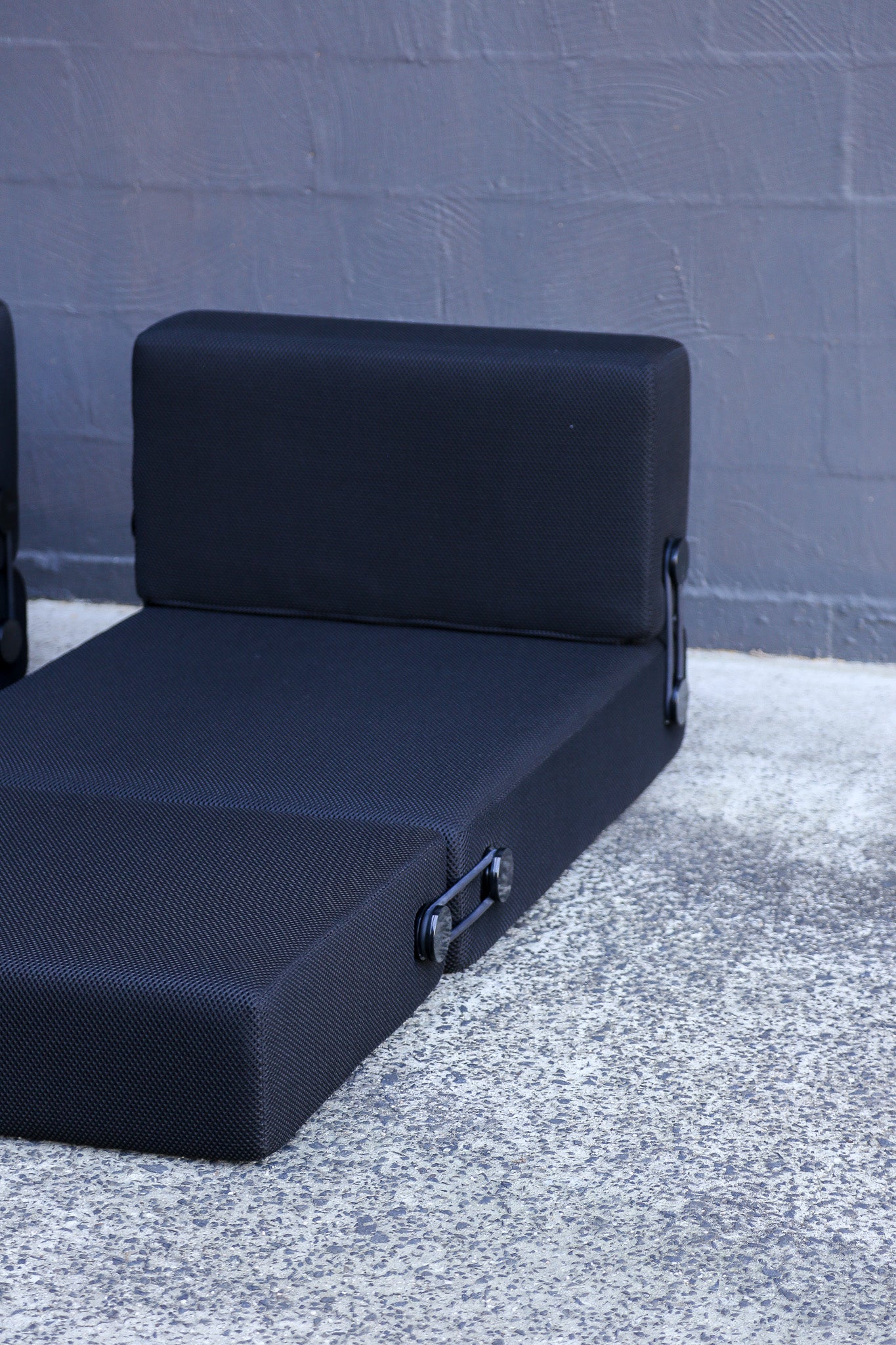 A black daybed with a textured fabric cover and metal side handles, positioned on a speckled concrete surface against a gray brick wall.