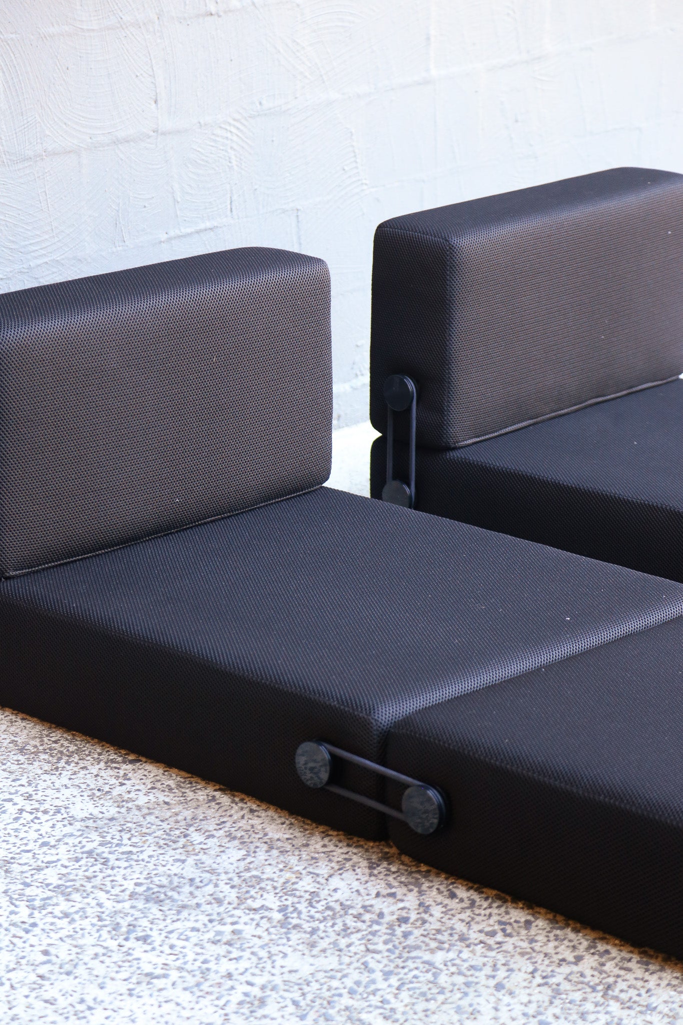 Black modular eating cushions with a textured fabric surface, shown against a white brick wall and speckled concrete floor.