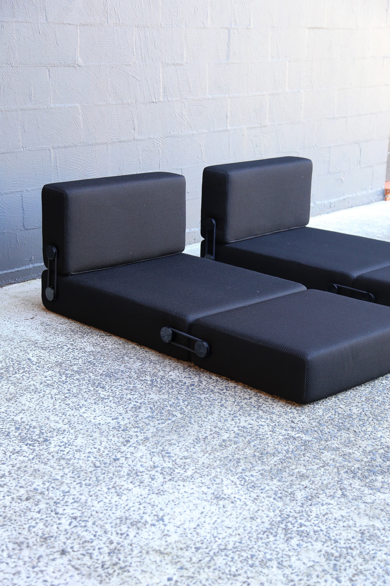 A black modular daybed with a high backrest, shown folded to reveal its storage compartment on a speckled concrete floor against a light gray brick wall.