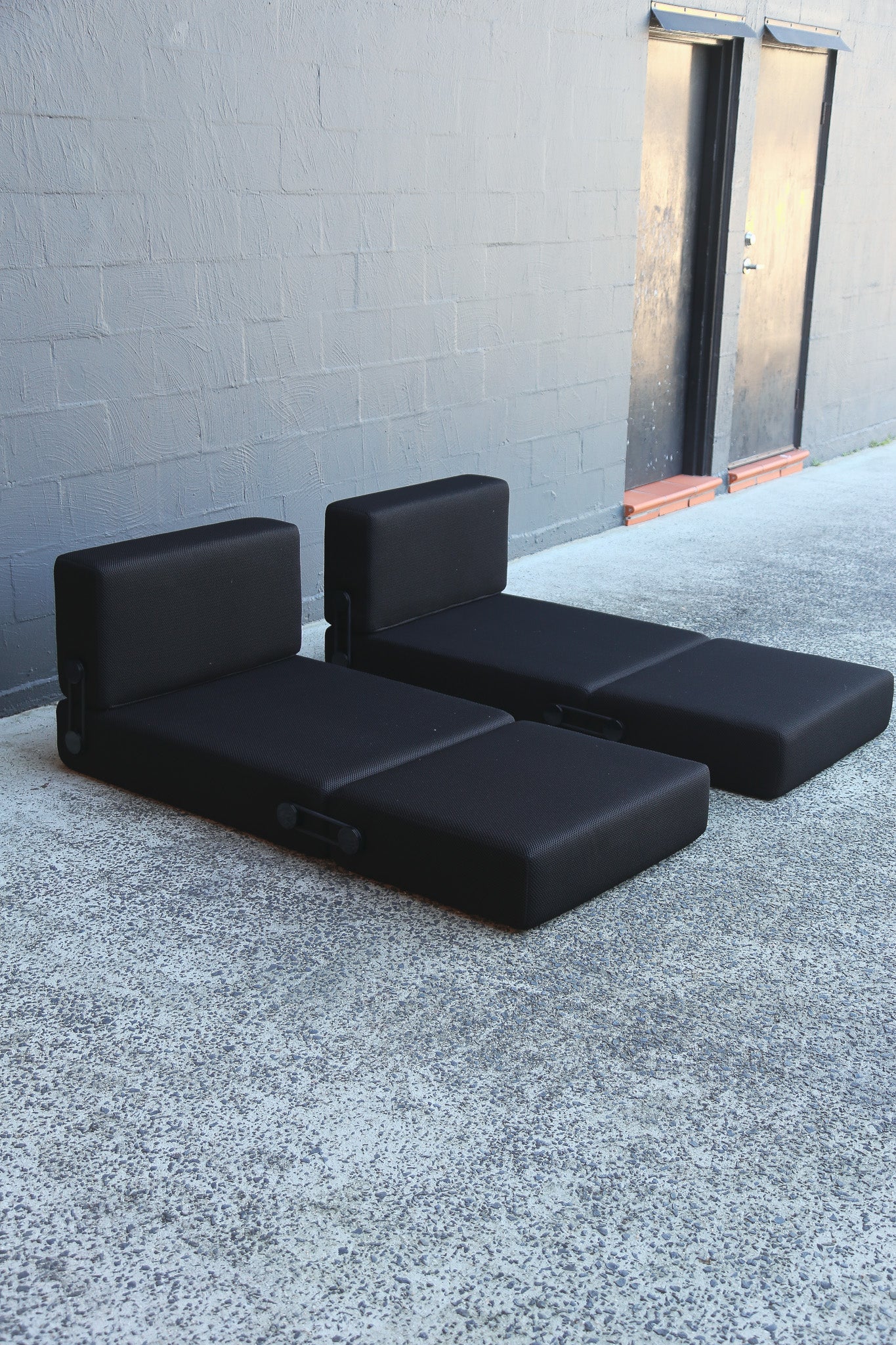 Two black daybeds with matching back cushions, positioned on a gray concrete surface against a gray brick wall.