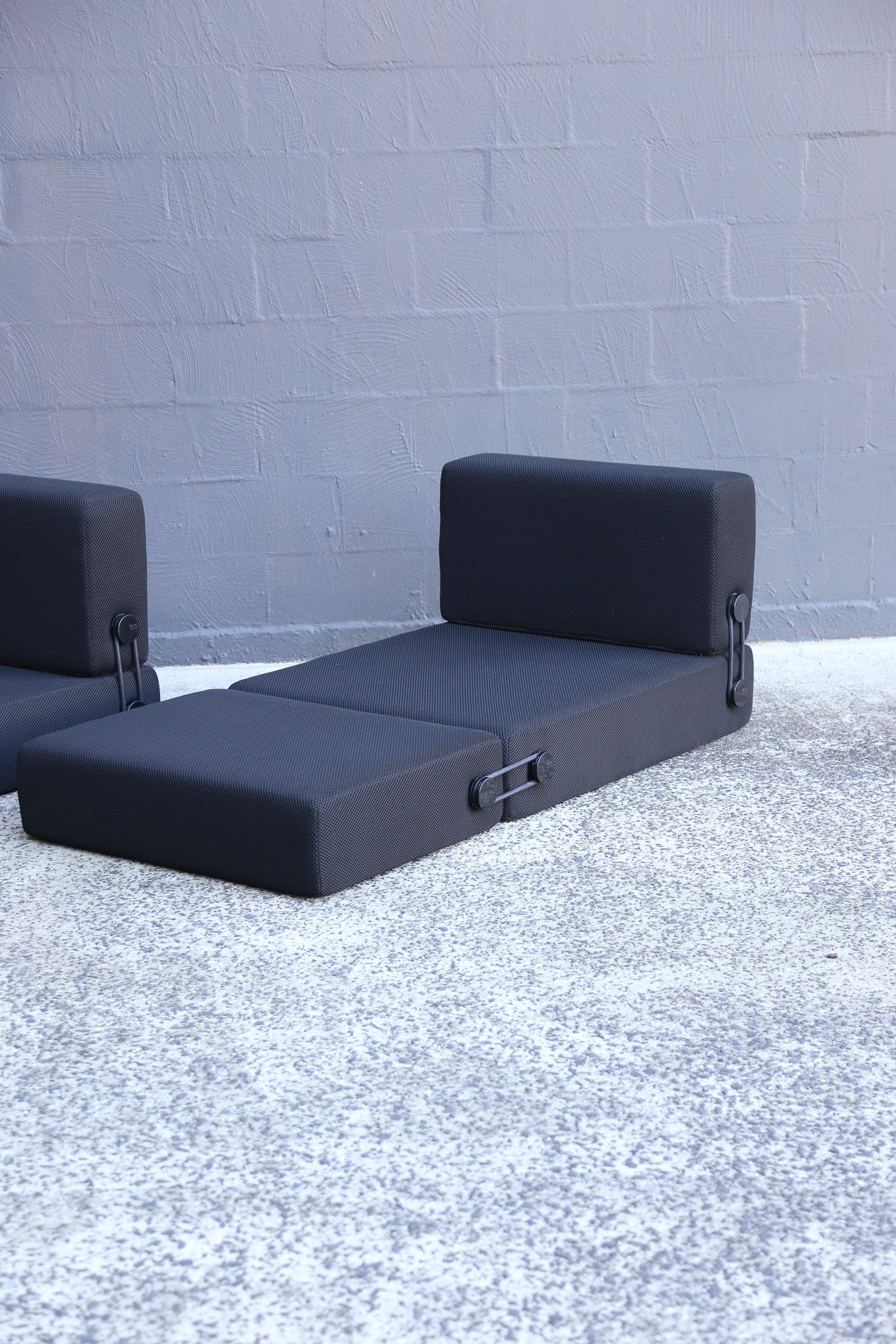 A dark gray modular daybed with a low backrest and side-release hardware, shown on a light gray concrete surface against a gray brick wall.