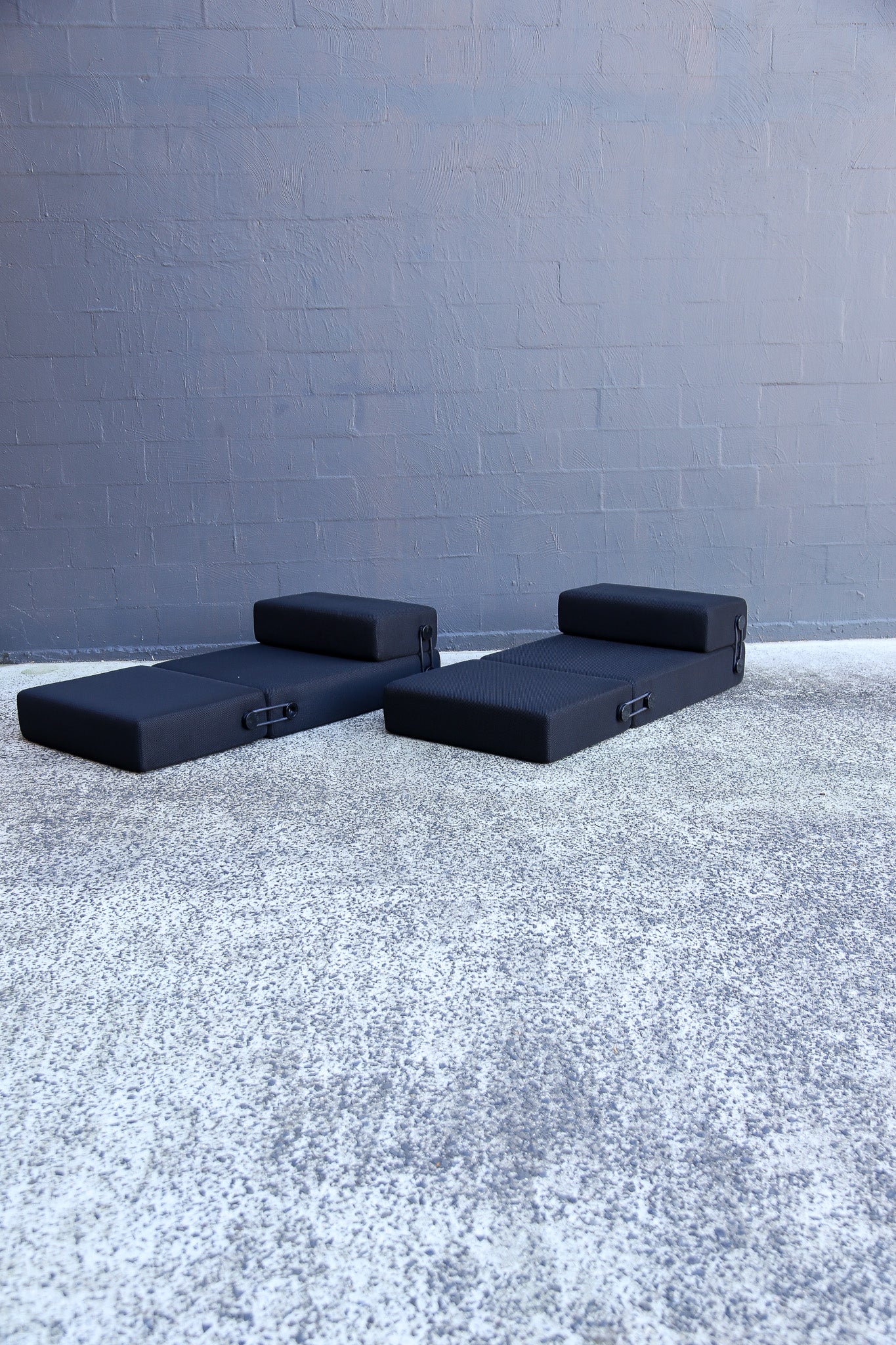Two black daybeds with integrated headrests and side storage compartments, placed on a textured concrete floor against a gray brick wall.
