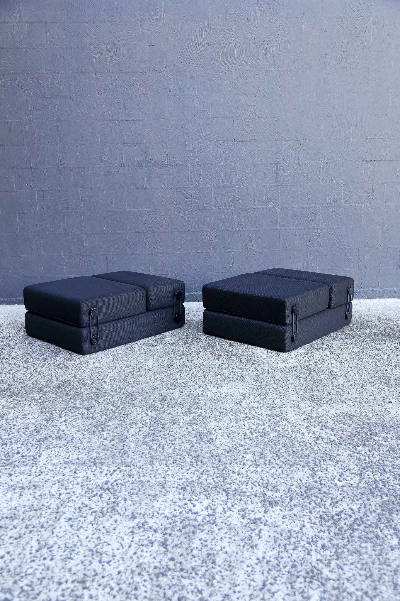 Two black rectangular modular sofa or floor cushions placed side by side on a textured gray floor against a gray brick wall.