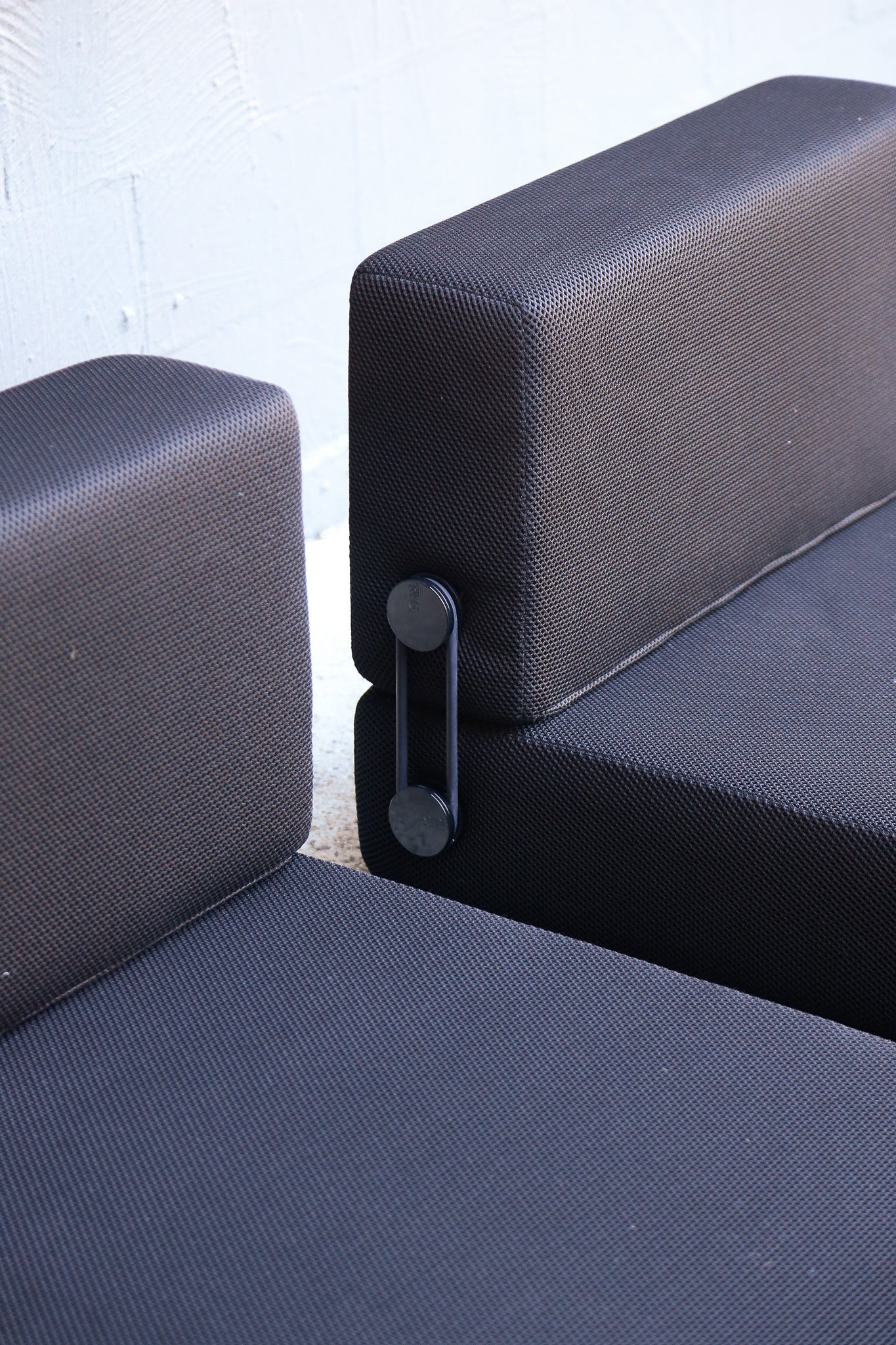 Close-up of a modular sofa with dark textured fabric cushions and a black metal tension-adjustment bracket between the armrests, set against a light off-white brick wall.