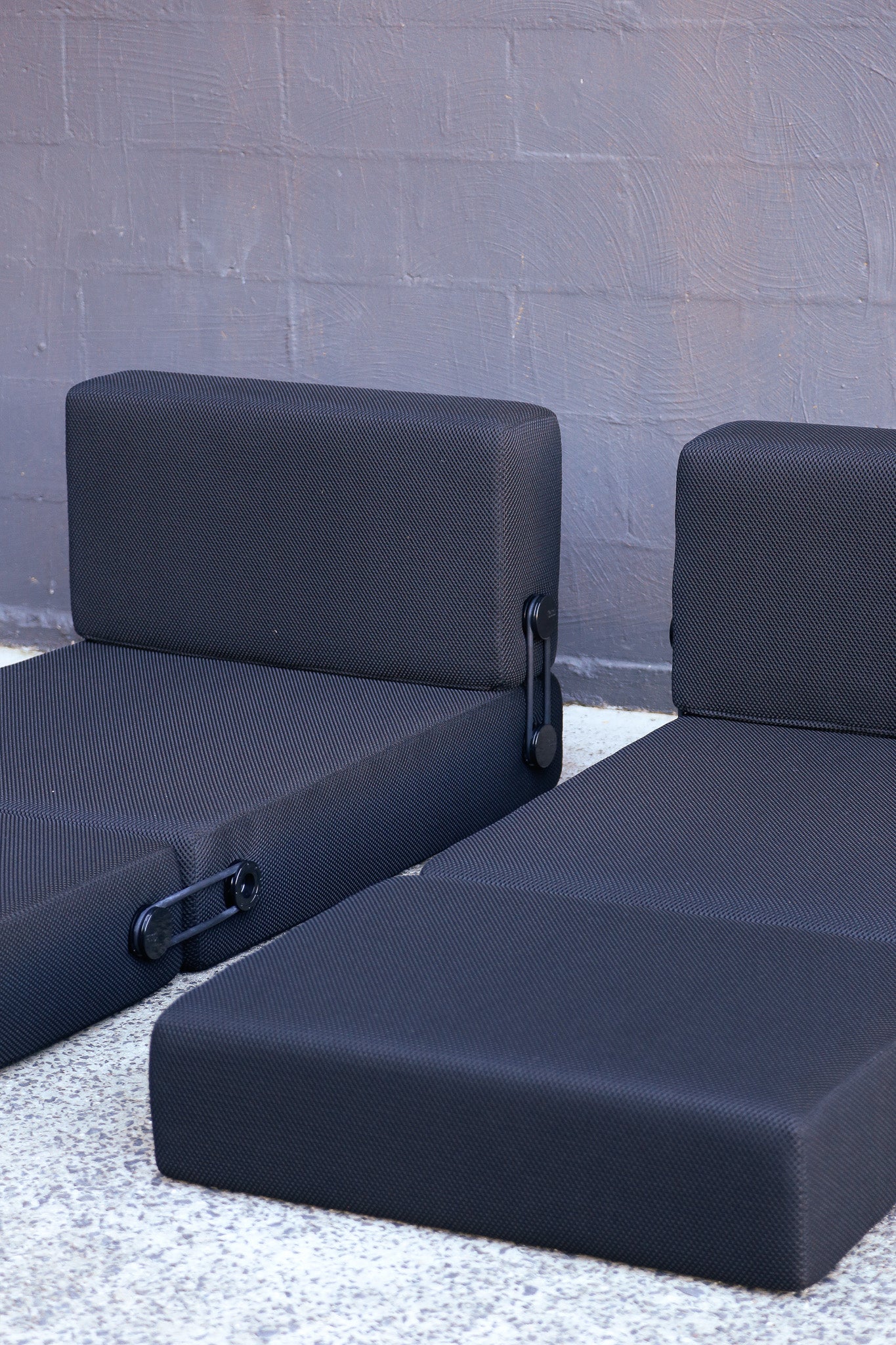 Two modular patio seating cushions in dark charcoal gray fabric, shown against a gray brick wall and light concrete ground.
