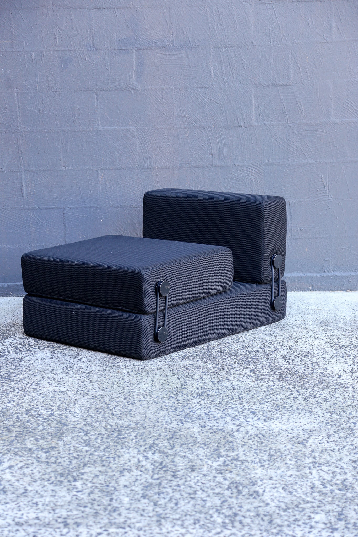 A black modular sofa cushion set consisting of a seat cushion, a backrest cushion, and a base frame, shown on a textured gray concrete floor against a gray painted brick wall.