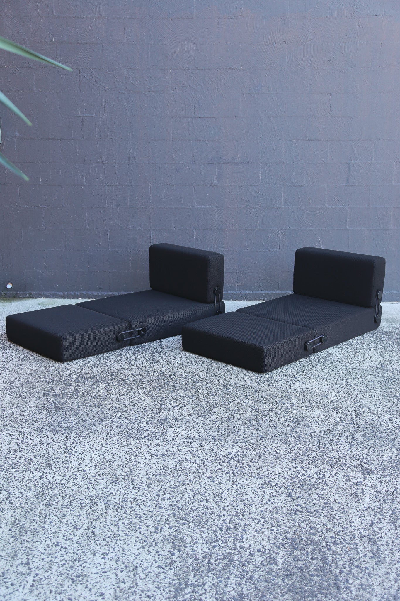 Two black modular daybeds with integrated headrests, positioned on a textured gray concrete surface against a dark gray brick wall.