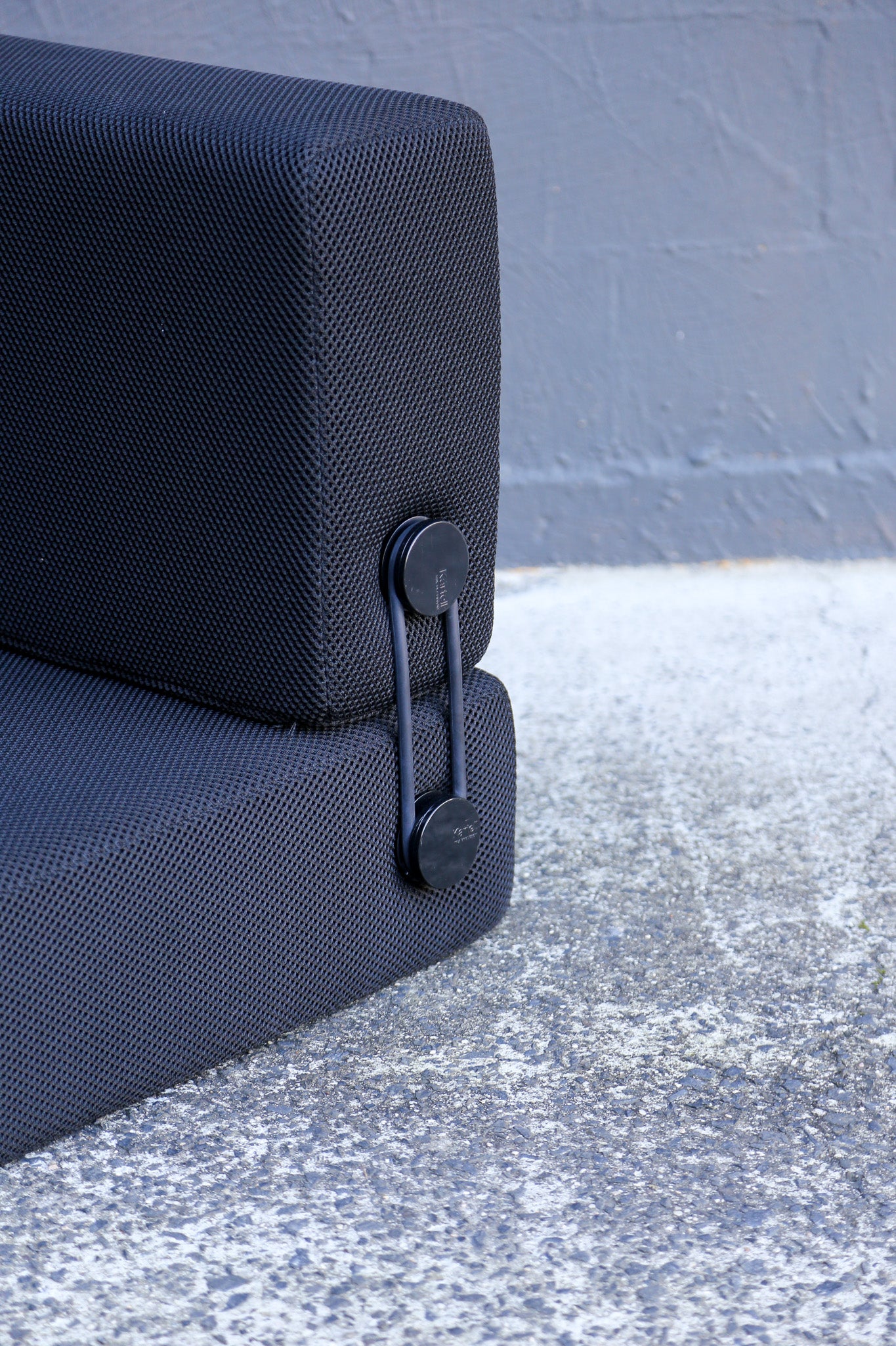 Close-up of a dark textured outdoor sofa cushion with a black metal safety latch, set against a gray concrete ground and wall.