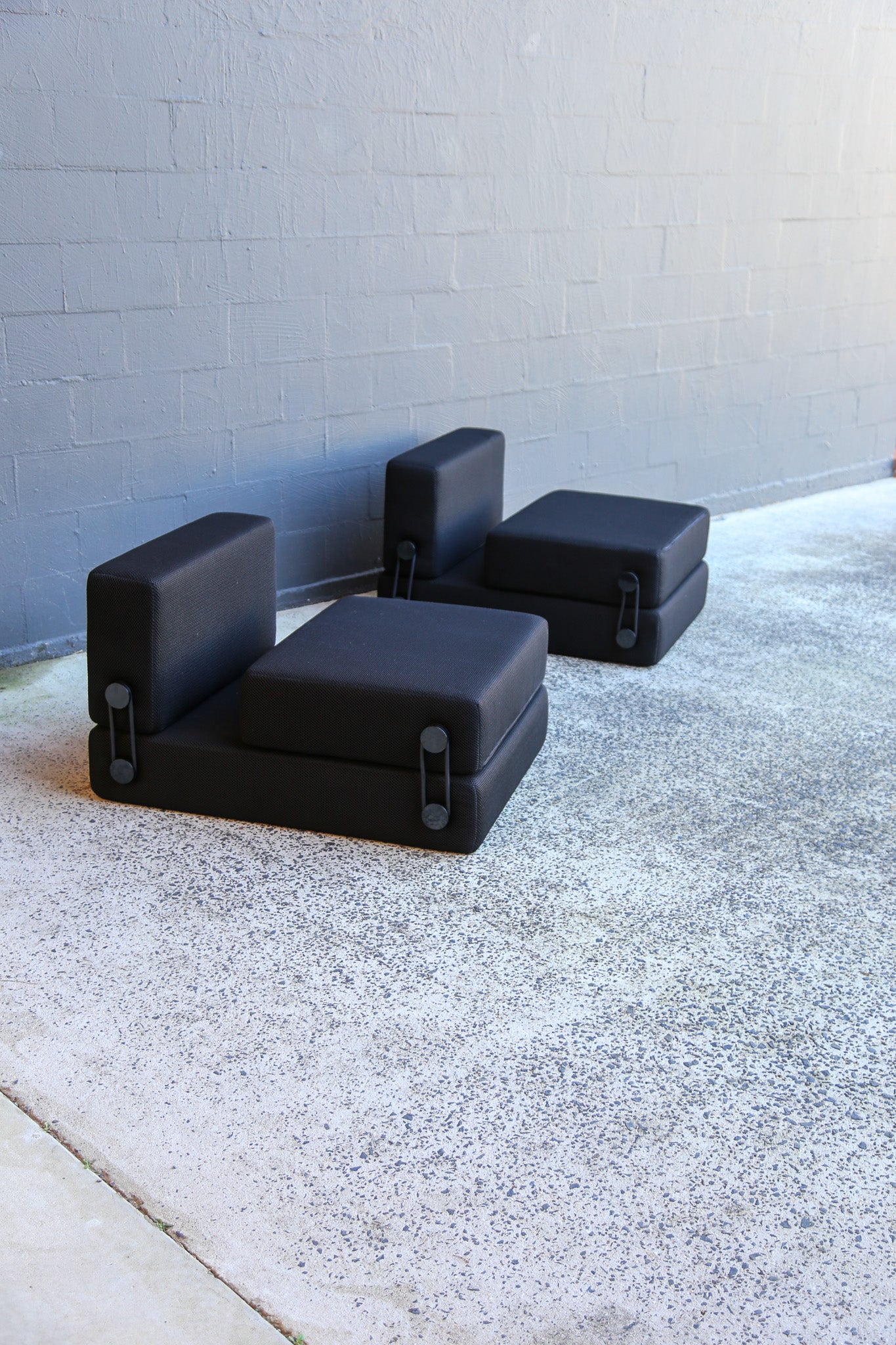 Trix modular black outdoor armchairs with integrated ottomans, arranged on a concrete patio against a gray brick wall.