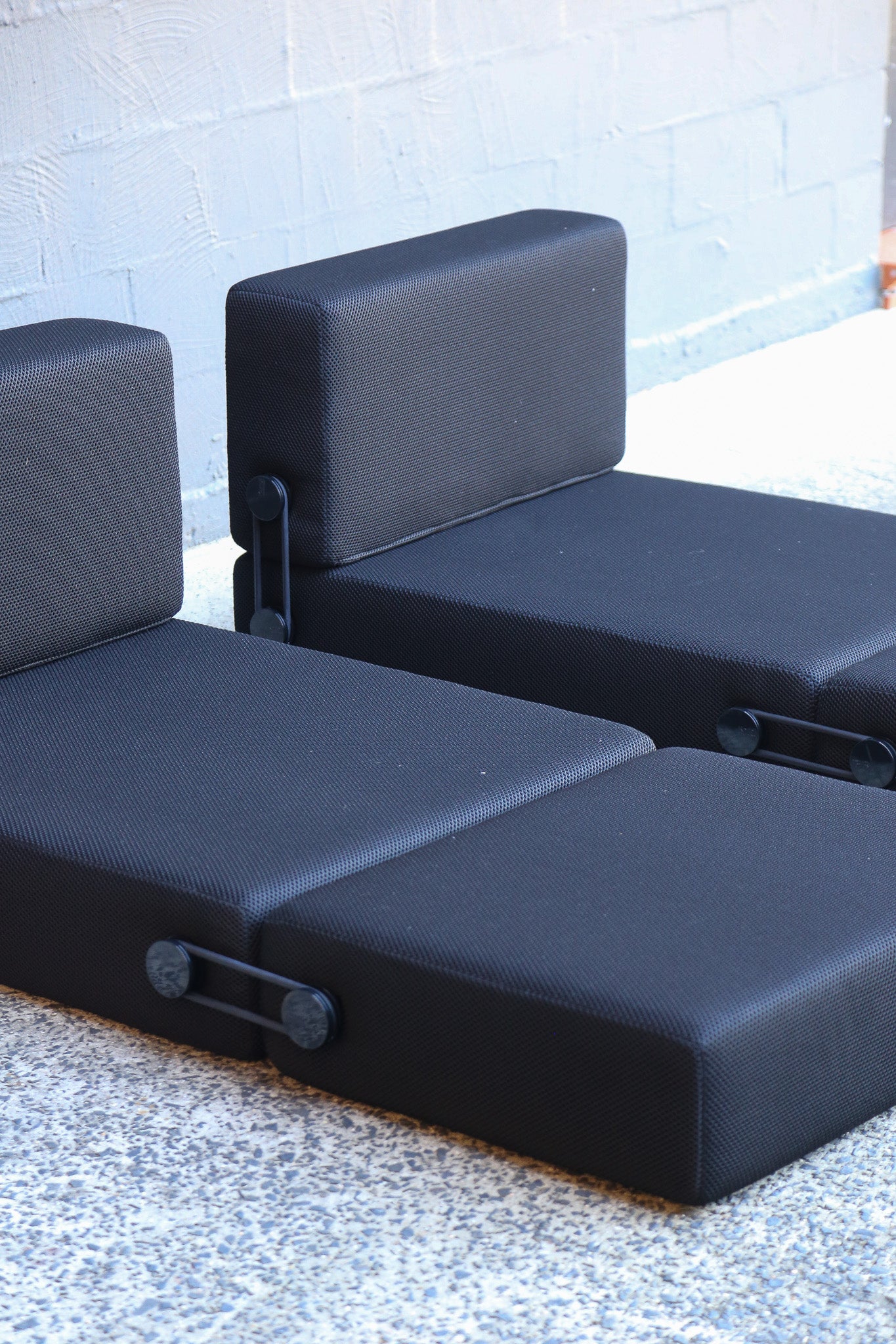 Two modular outdoor patio seating modules with black textured fabric cushions, shown on a speckled concrete floor against a light gray brick wall.