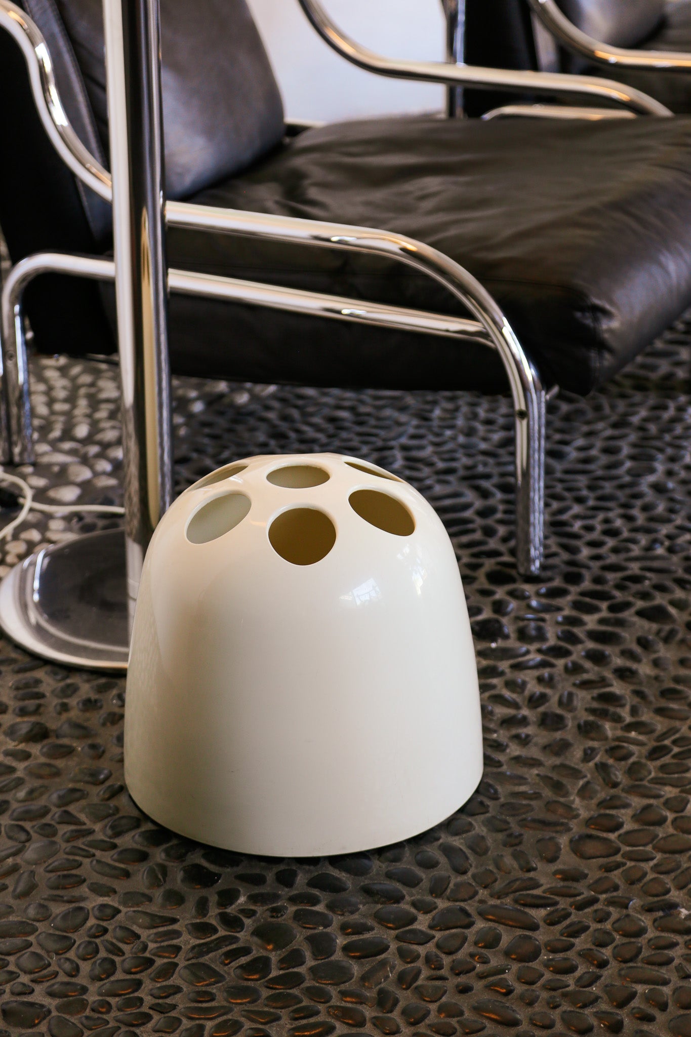 White umbrella stand with holes on a textured surface next to a black leather chair.