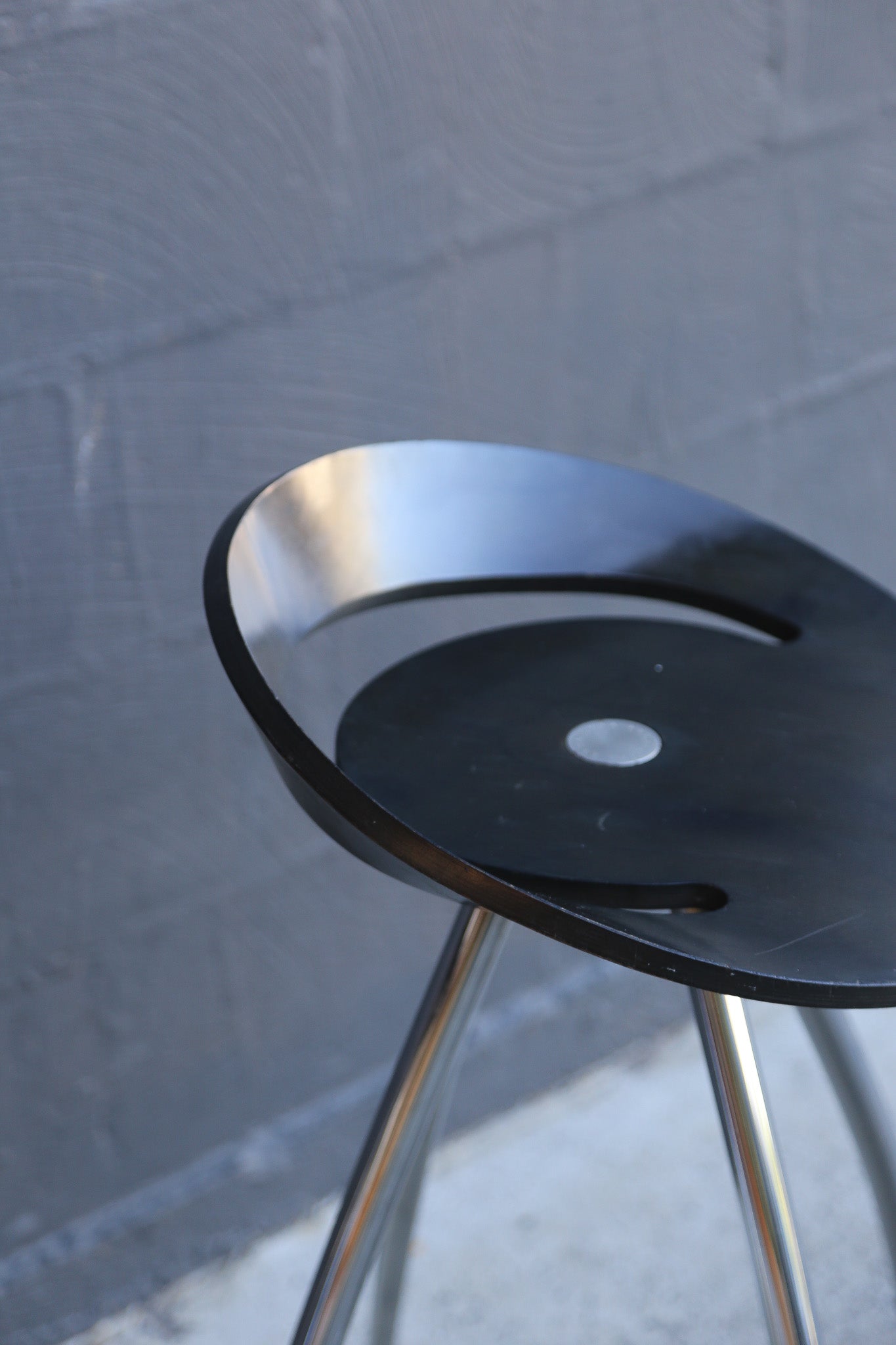 Modern stool with black seat and silver legs against a textured gray wall.