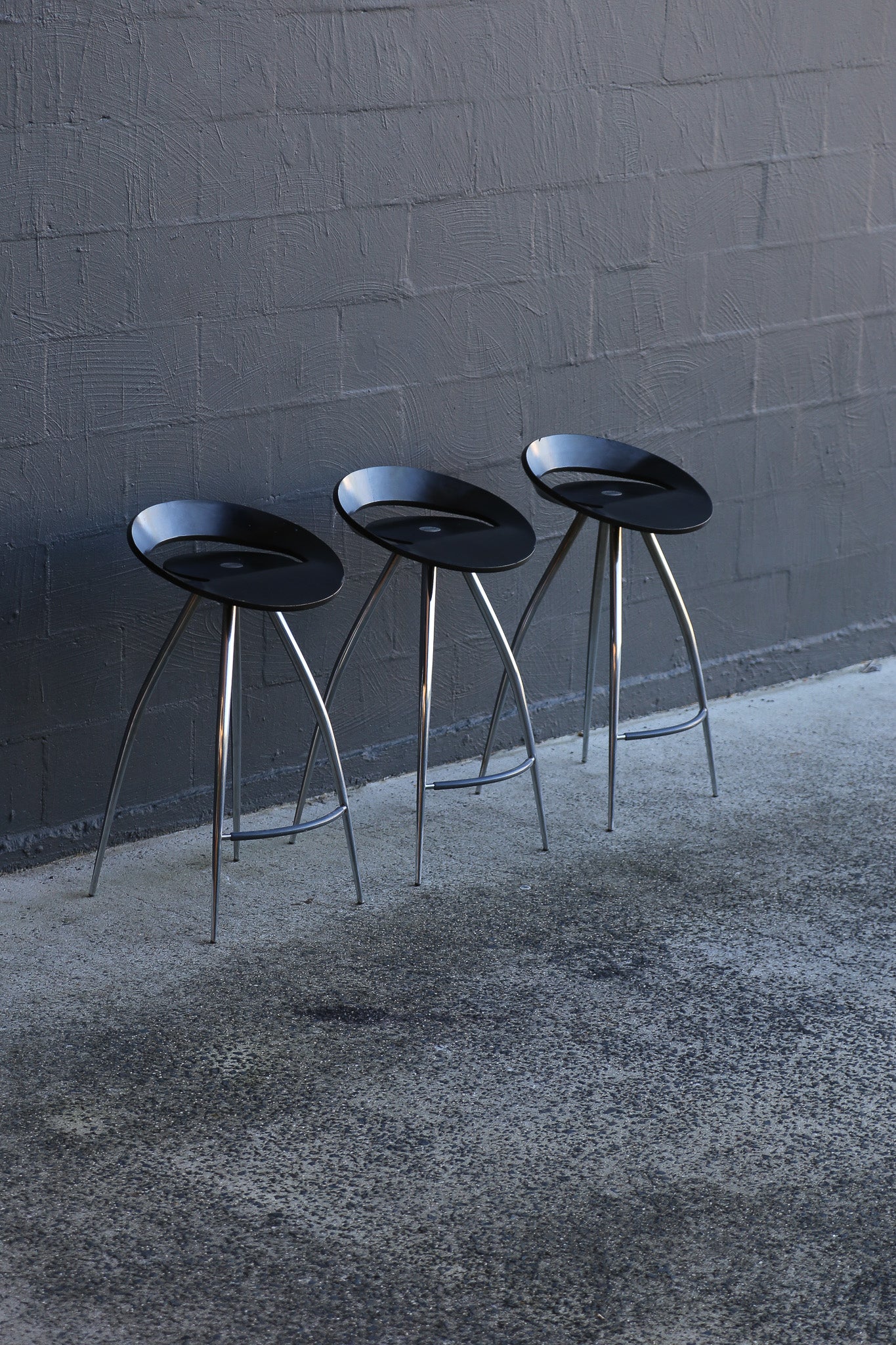 Three black bar stools with metal legs against a gray brick wall.