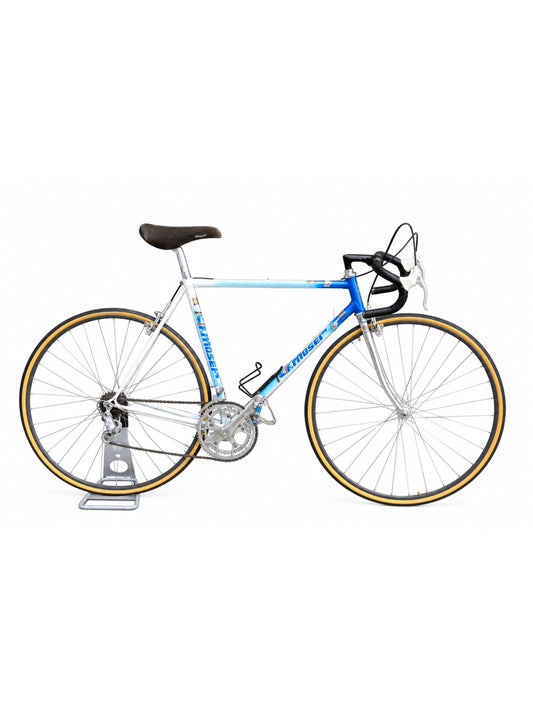 A vintage road bicycle with a blue and white frame, black saddle and handlebars, and tan sidewall tires, shown on a silver stand against a white background.